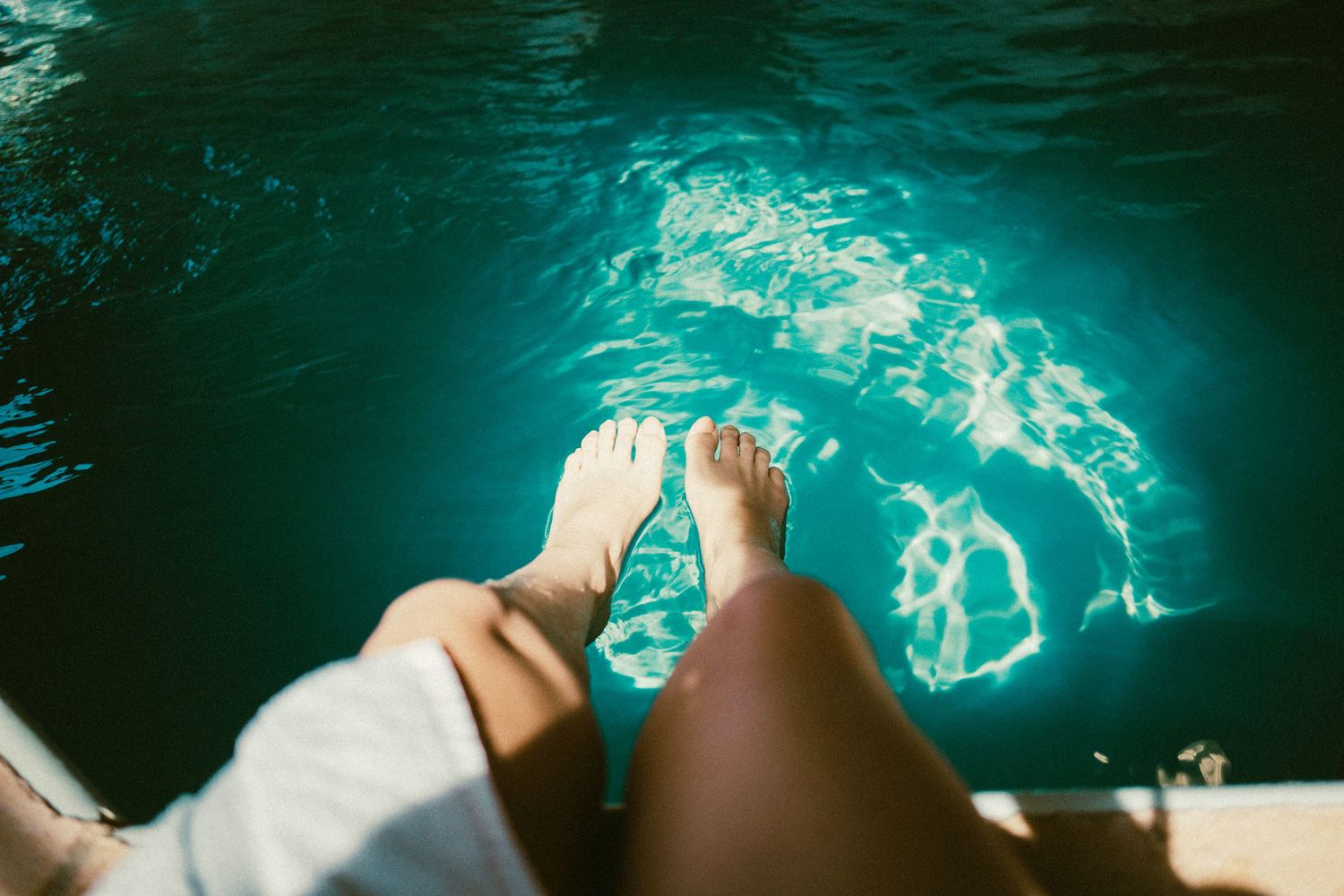 Person dangling bare feet over a shimmering turquoise swimming pool on a sunny day.