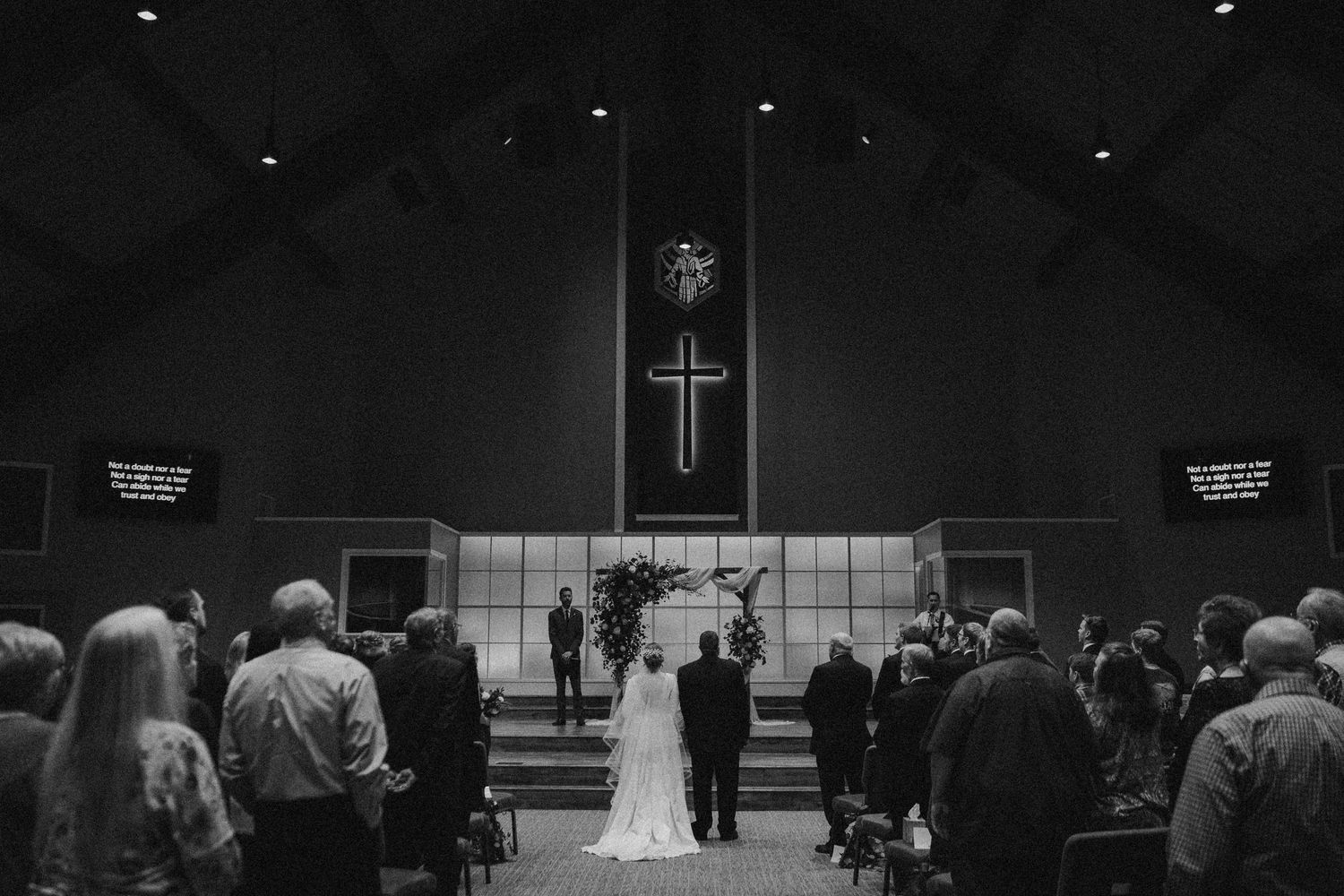 Black and white wedding ceremony photo inside a church with a cross, bride and groom at altar, guests standing in pews.