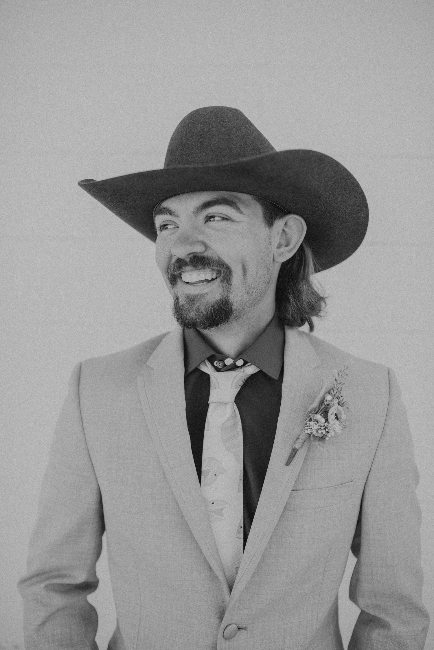 Smiling man in cowboy hat, light suit, black tie, and boutonniere poses outdoors in black and white photo.