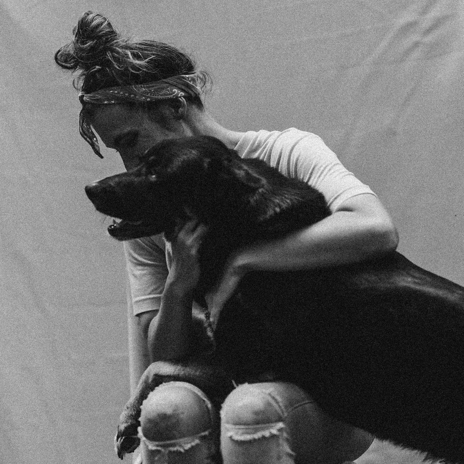 Black and white photo of a young woman hugging a large black dog, both sharing a tender moment against a light background.