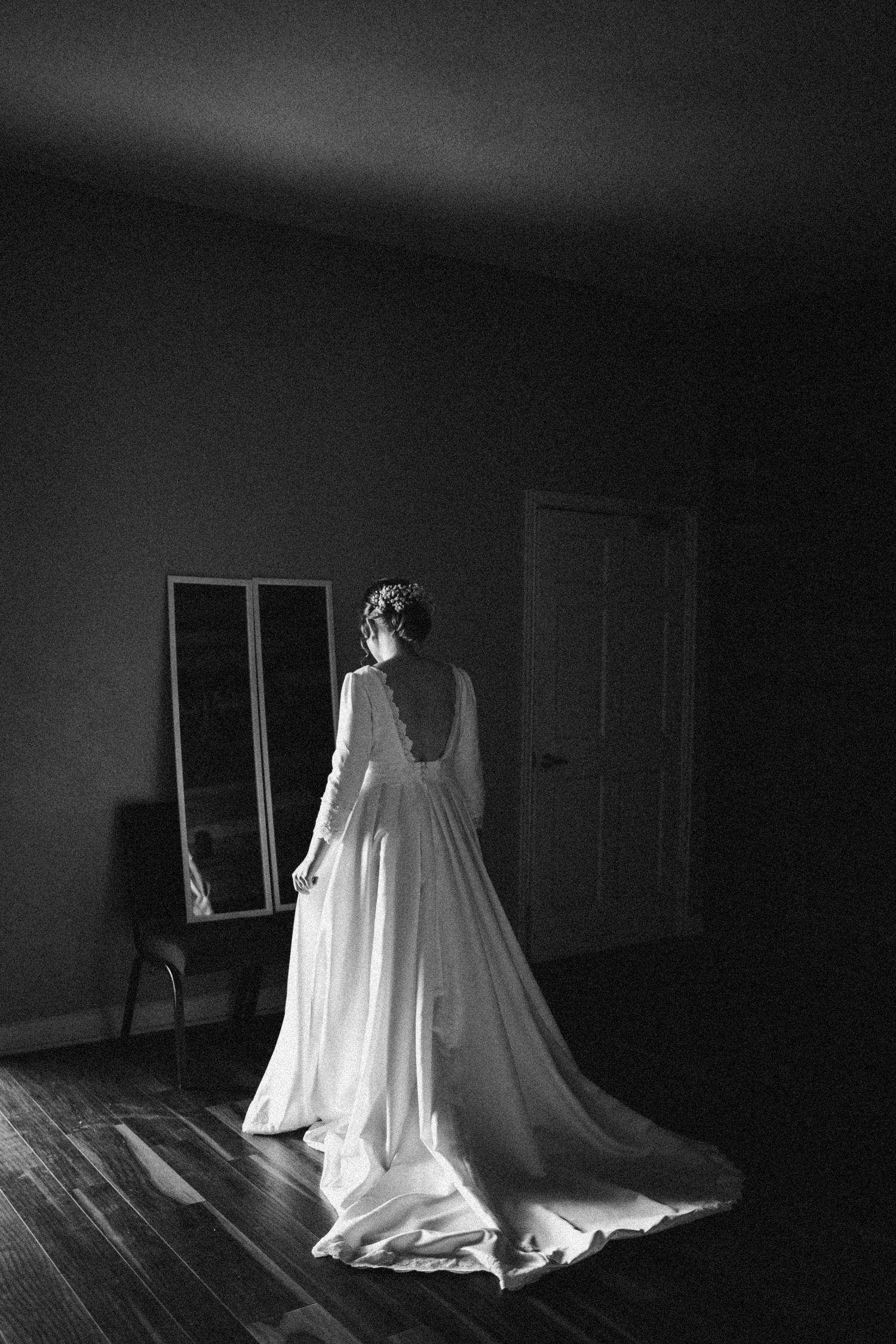 Bride in elegant backless long-sleeve wedding gown with train stands in dimly lit room beside mirror, black and white photo.