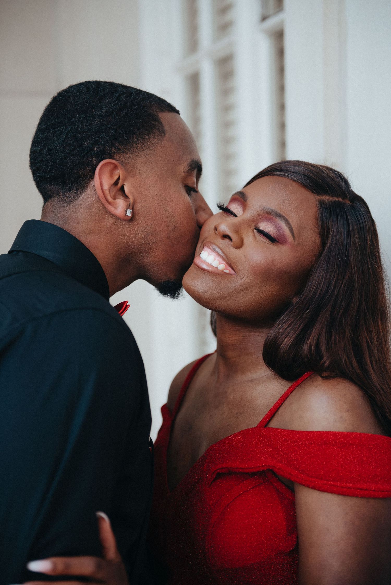 Joyful Black couple sharing a romantic kiss, woman in red off-shoulder dress smiling with eyes closed.