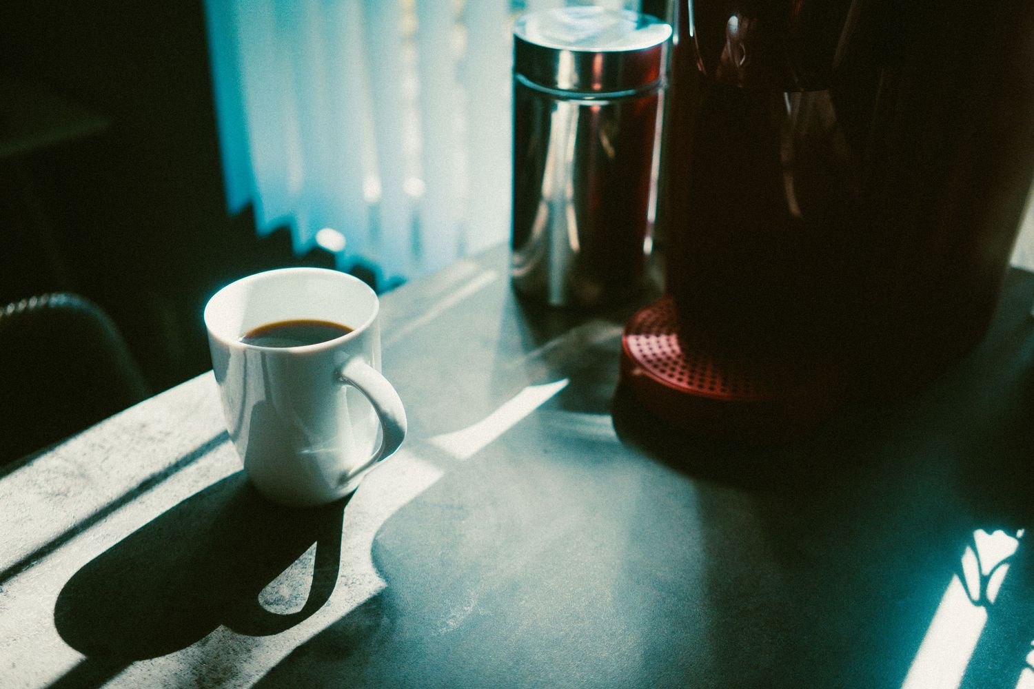 A white coffee mug sits on a dark countertop beside a coffee maker, bathed in warm morning sunlight streaming through blinds.