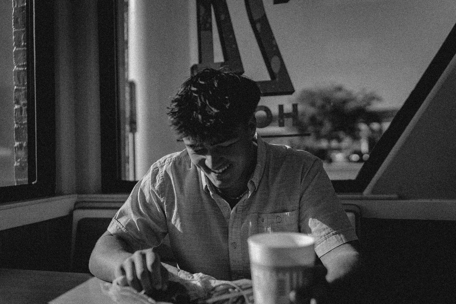 Black and white photo of a young man smiling while eating at a diner booth near a window.
