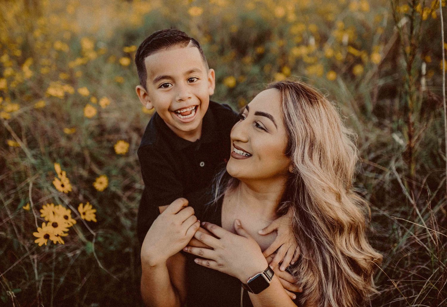 A joyful boy hugs a smiling woman in a sunny field of yellow wildflowers, capturing a warm mother-son moment.