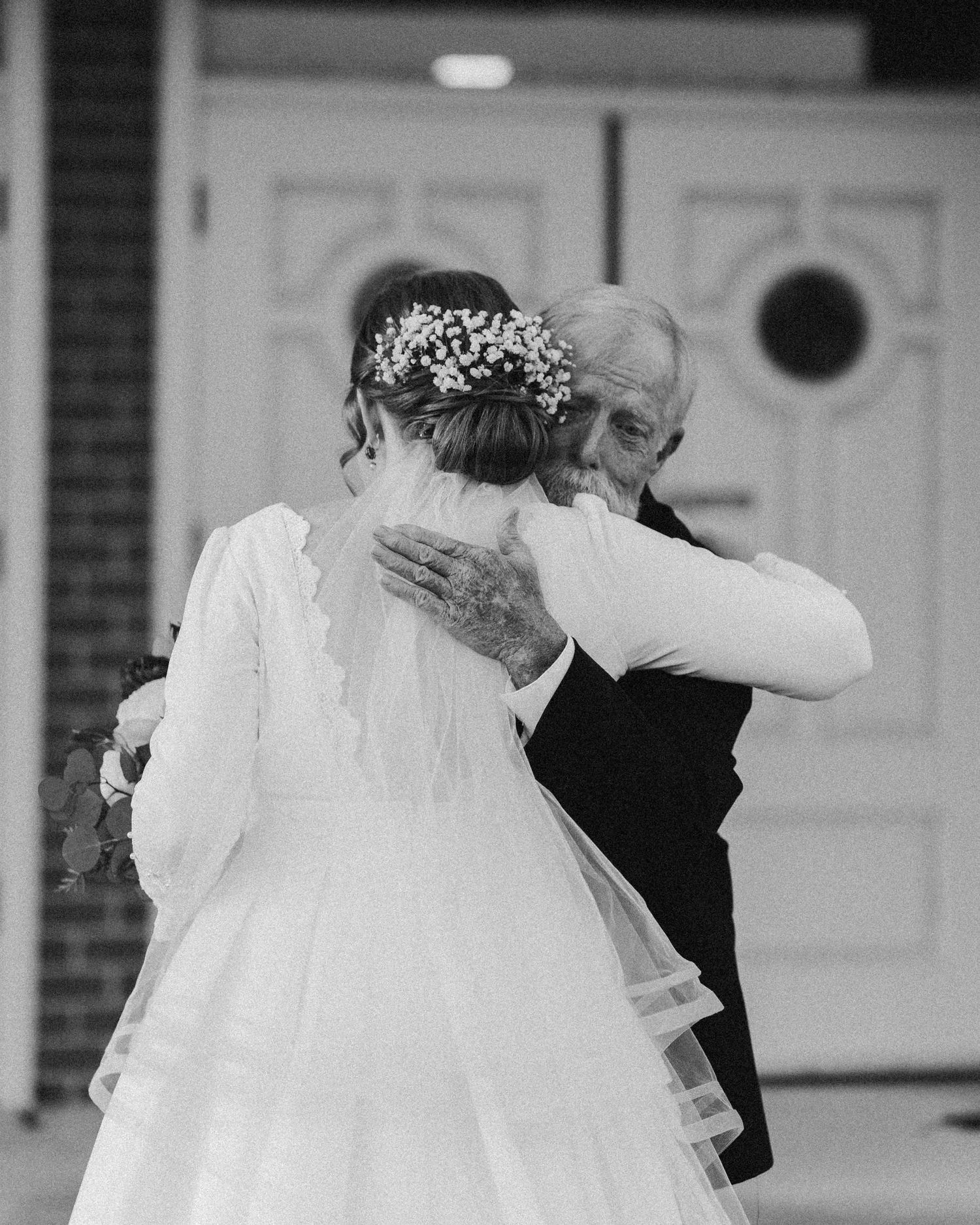 Black and white photo of a bride hugging an elderly man in a dark suit outside a white door.