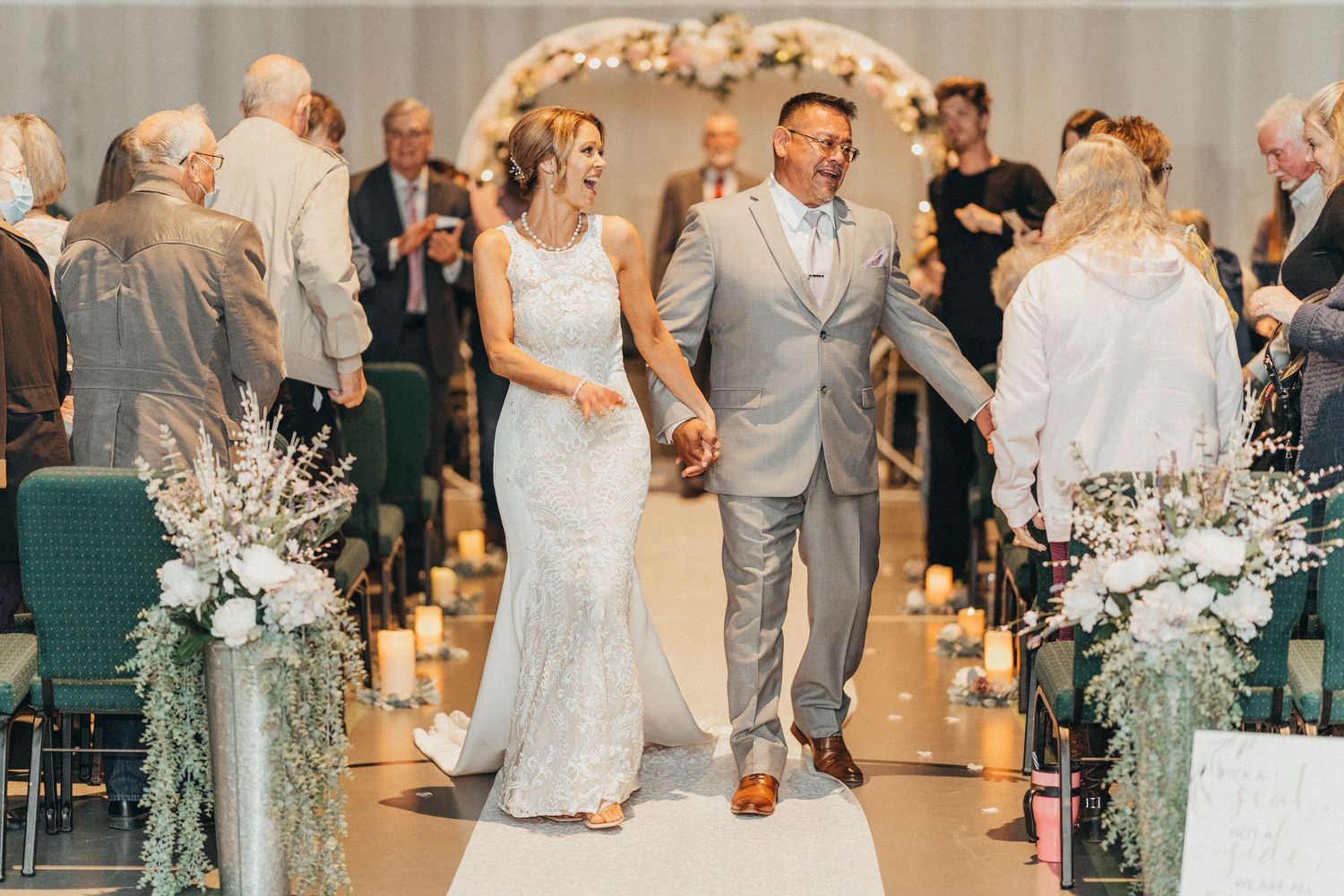 Joyful newlyweds walk down the aisle after wedding ceremony, surrounded by floral arrangements and candles.
