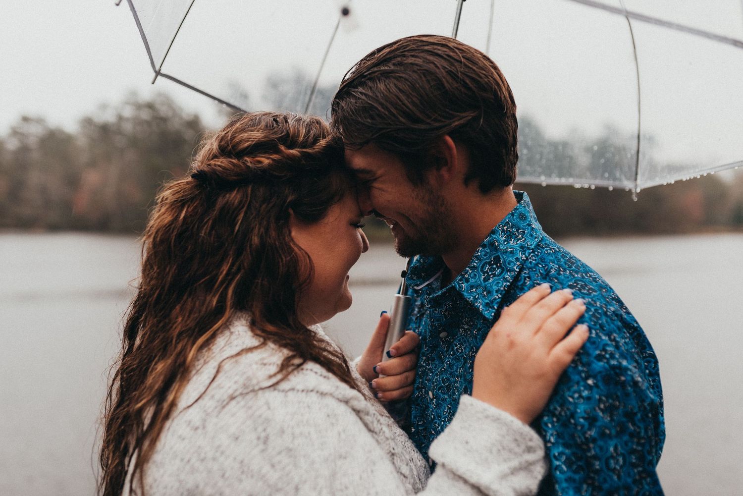 Couple smiling and touching foreheads under a clear umbrella on a rainy day near a lake.
