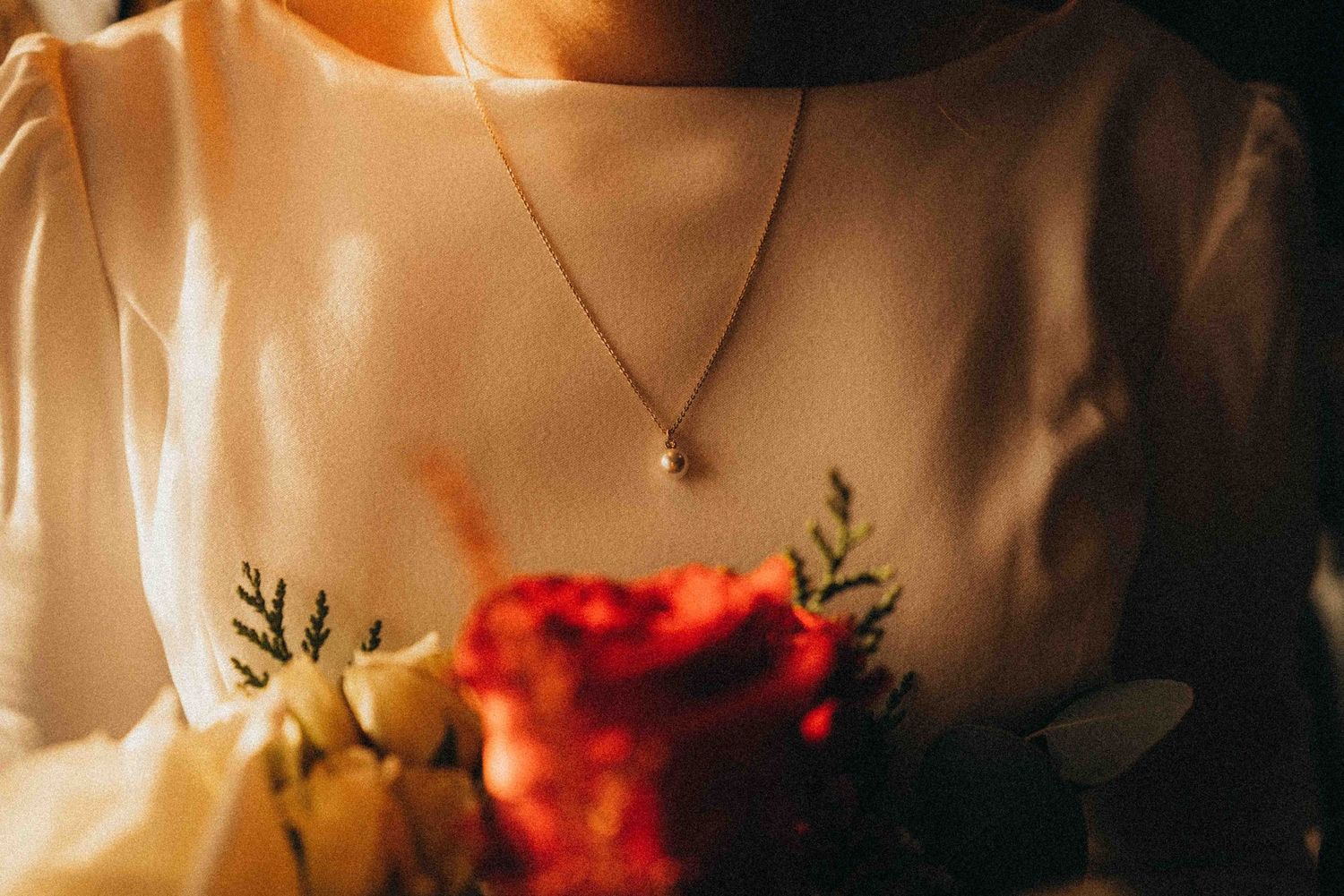 Close-up of a bride holding a red and white bouquet, wearing a pearl necklace and white embroidered dress.