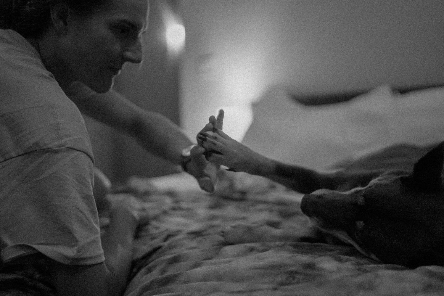 Black and white photo of a woman on a bed gently touching paws with a dark-colored dog in soft light.