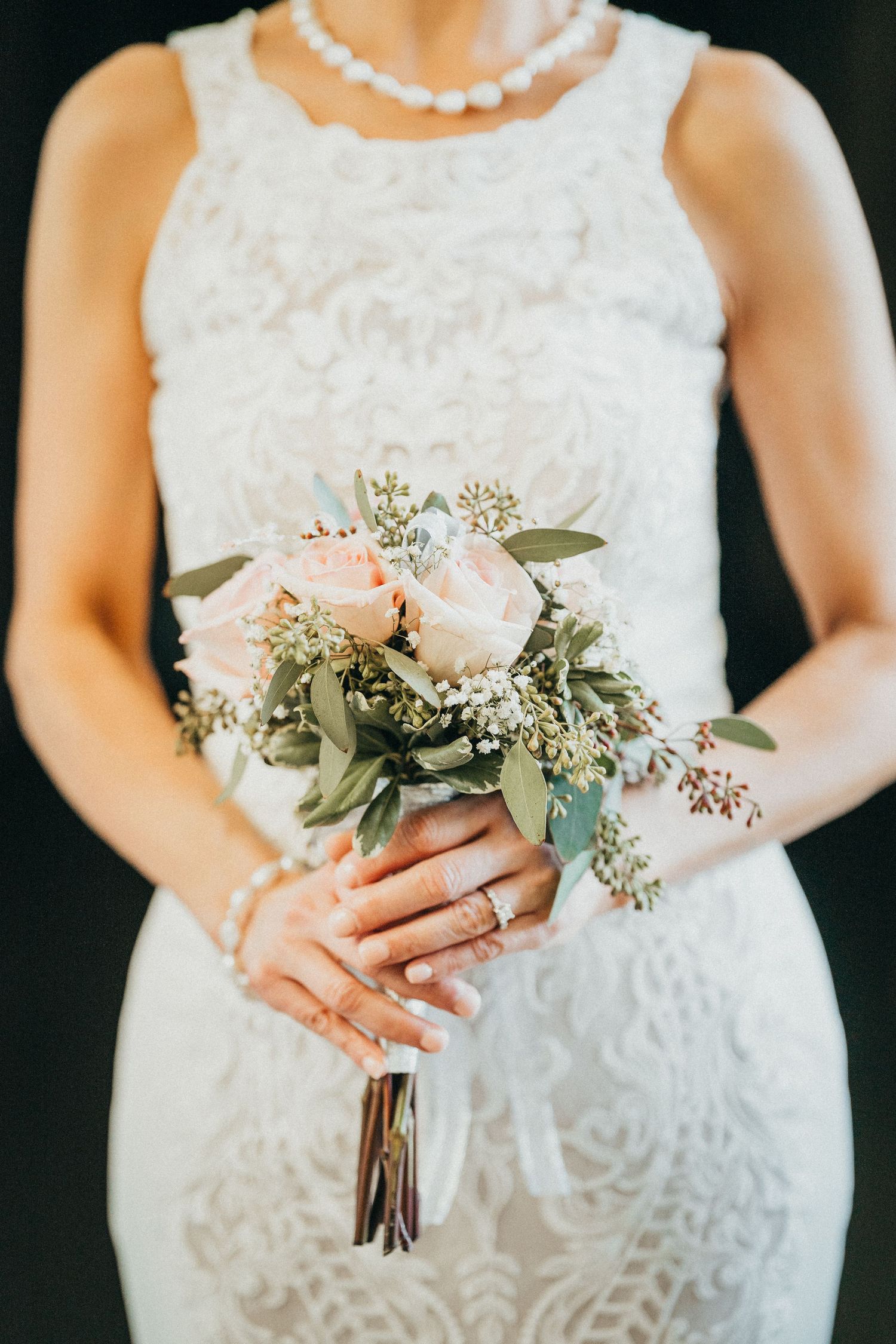 Bride in white lace dress holding a small bouquet of blush roses and greenery, wearing pearl jewelry.