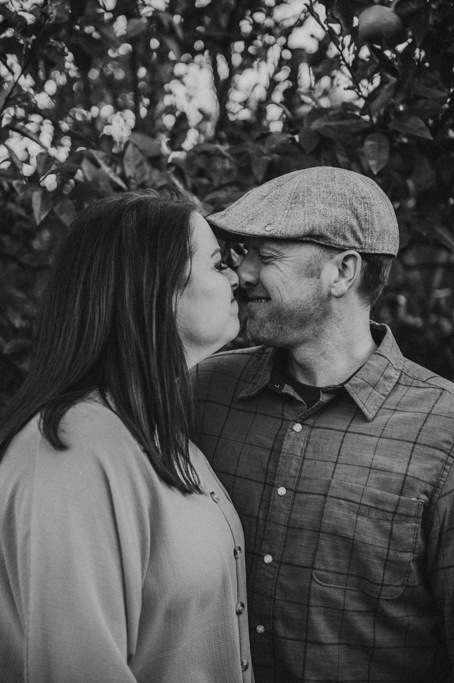 Black and white photo of a couple about to kiss outdoors, man wearing a flat cap and plaid shirt, woman in a light jacket.