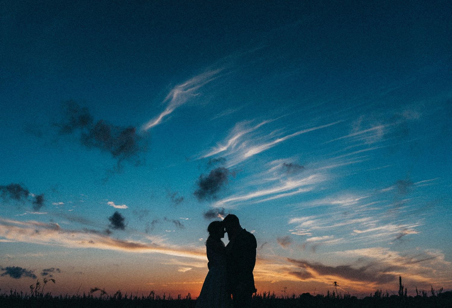 Romantic silhouette of wedding couple embracing against stunning blue and orange sunset sky.