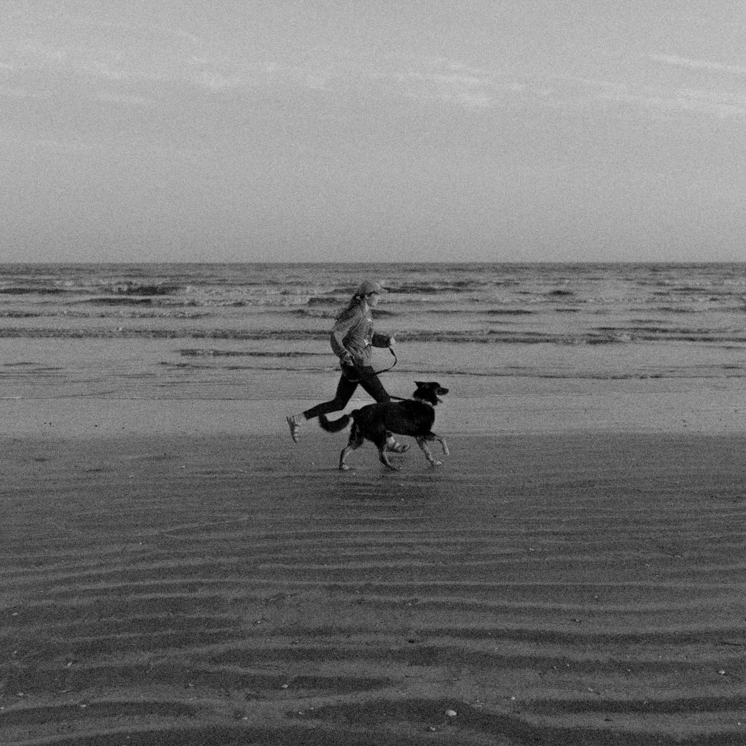 Person running with two dogs on a wide sandy beach in black and white photography.