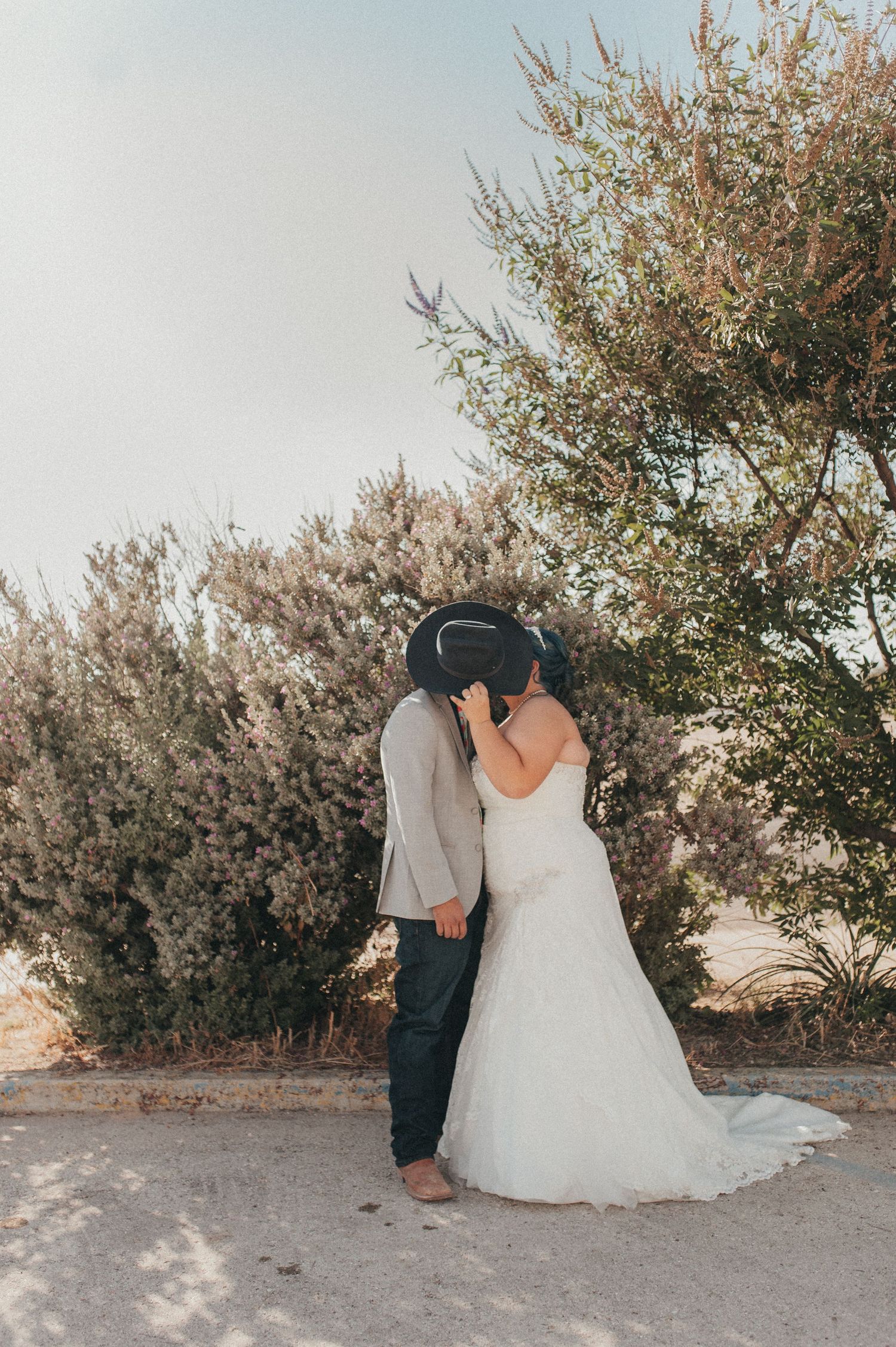 A bride and groom share a kiss outdoors on their wedding day, with lush green shrubs and a clear sky behind them.