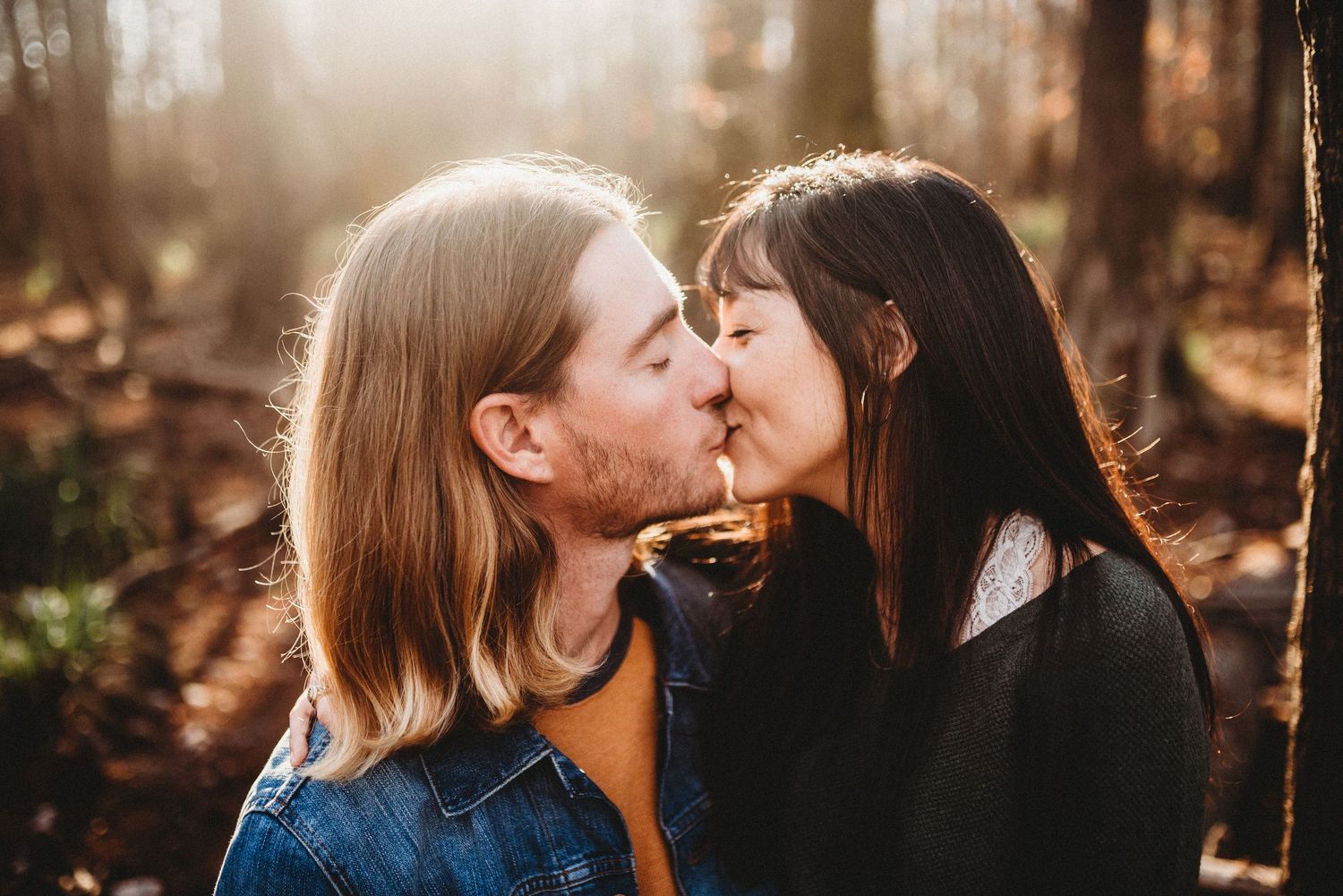 A couple shares a romantic kiss in a sunlit forest, with warm golden light filtering through the trees behind them.