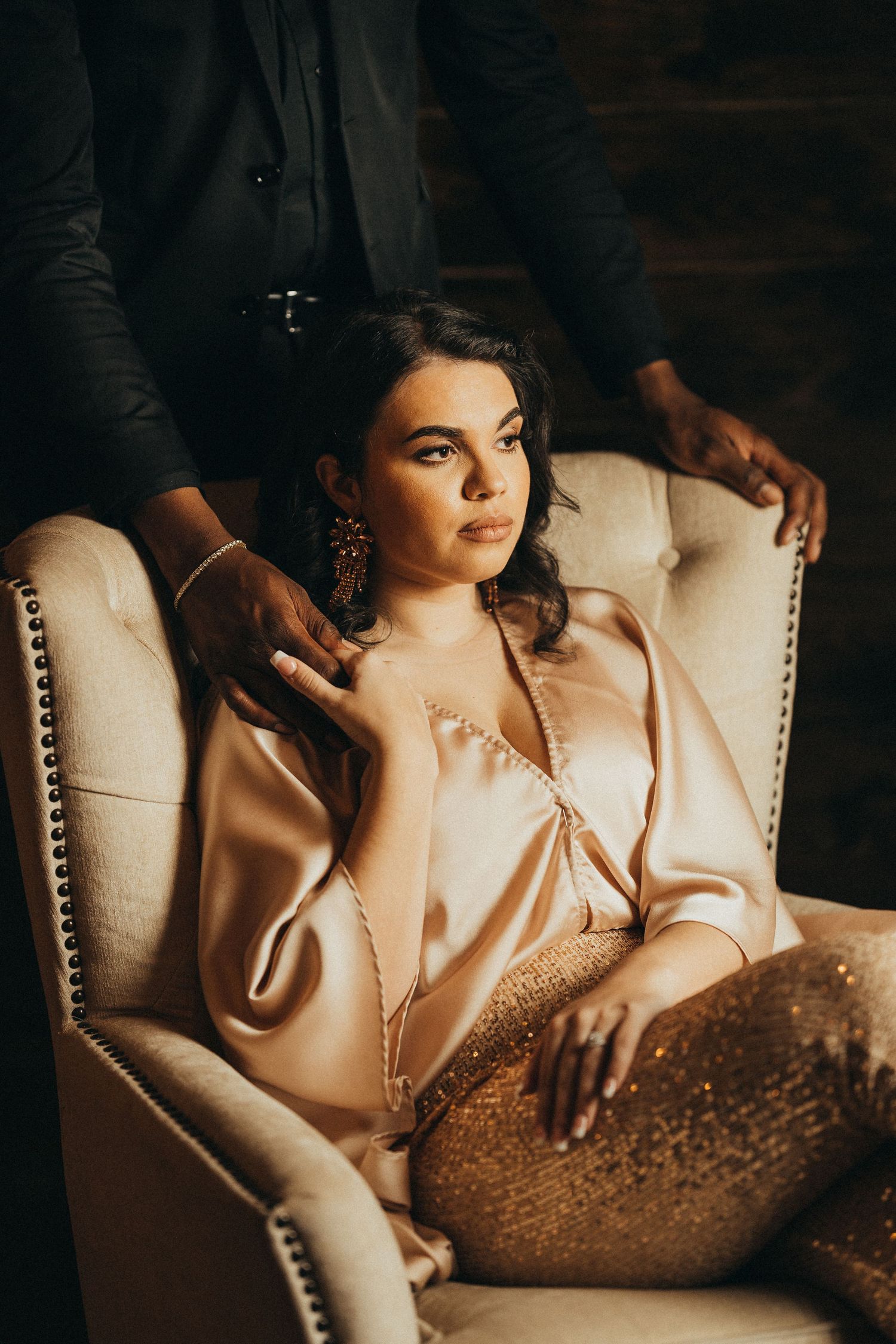 Woman in satin blouse and gold sequin pants seated in tufted chair with man in dark suit standing behind.