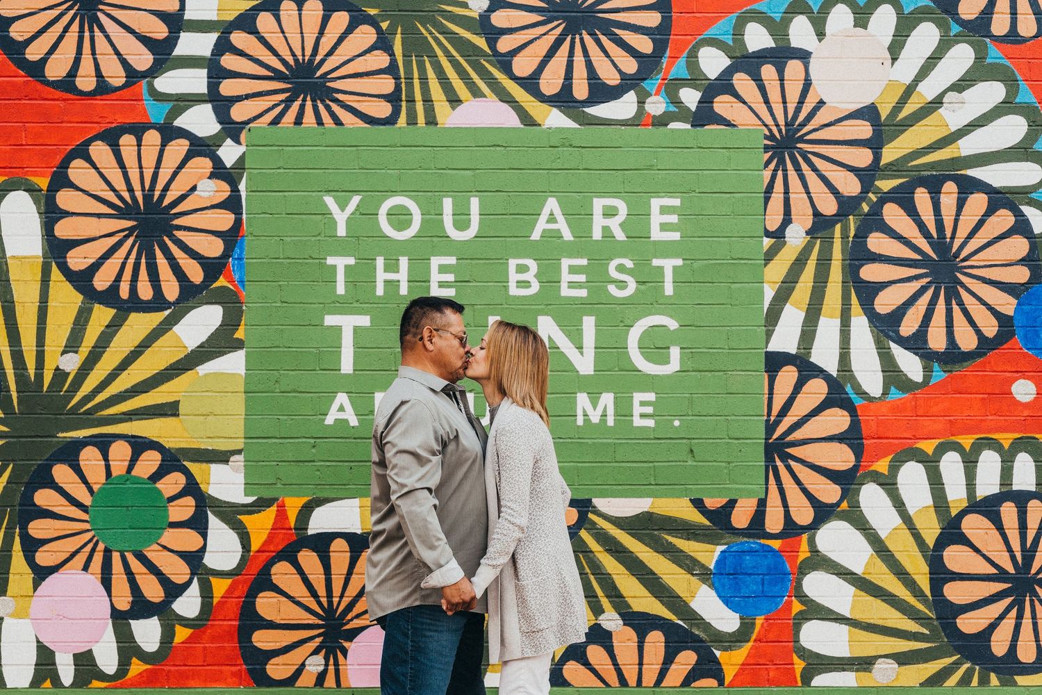 Couple sharing a kiss in front of a colorful floral mural with an inspiring love quote in green.