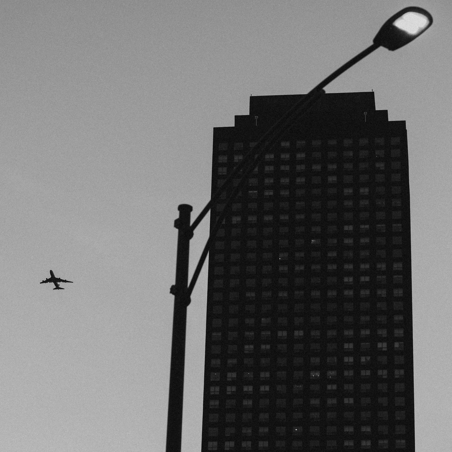 Black and white photo of a tall city skyscraper with a street lamp and airplane flying in the overcast sky.