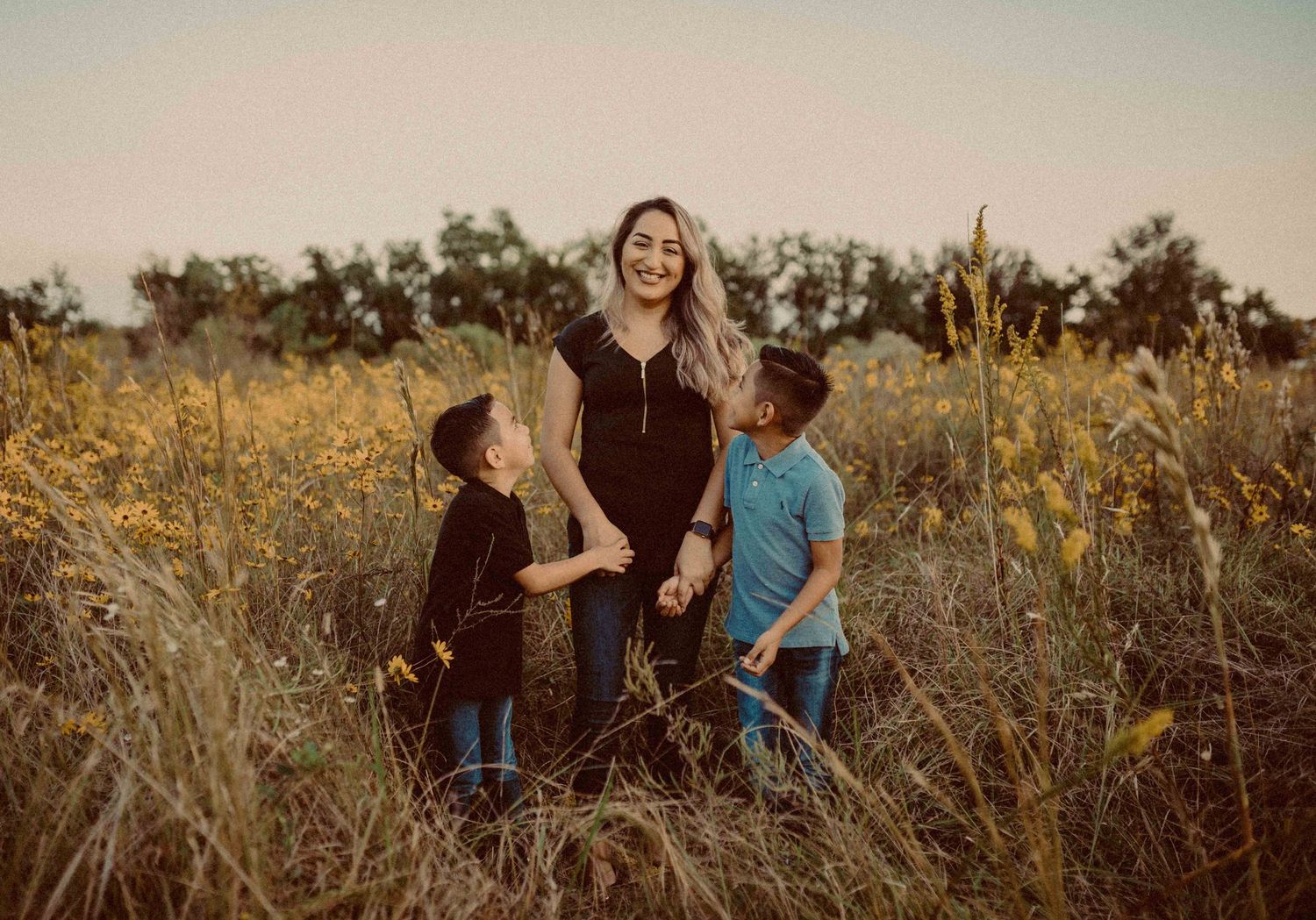 A mother smiles warmly with her two young sons in a golden field at sunset.
