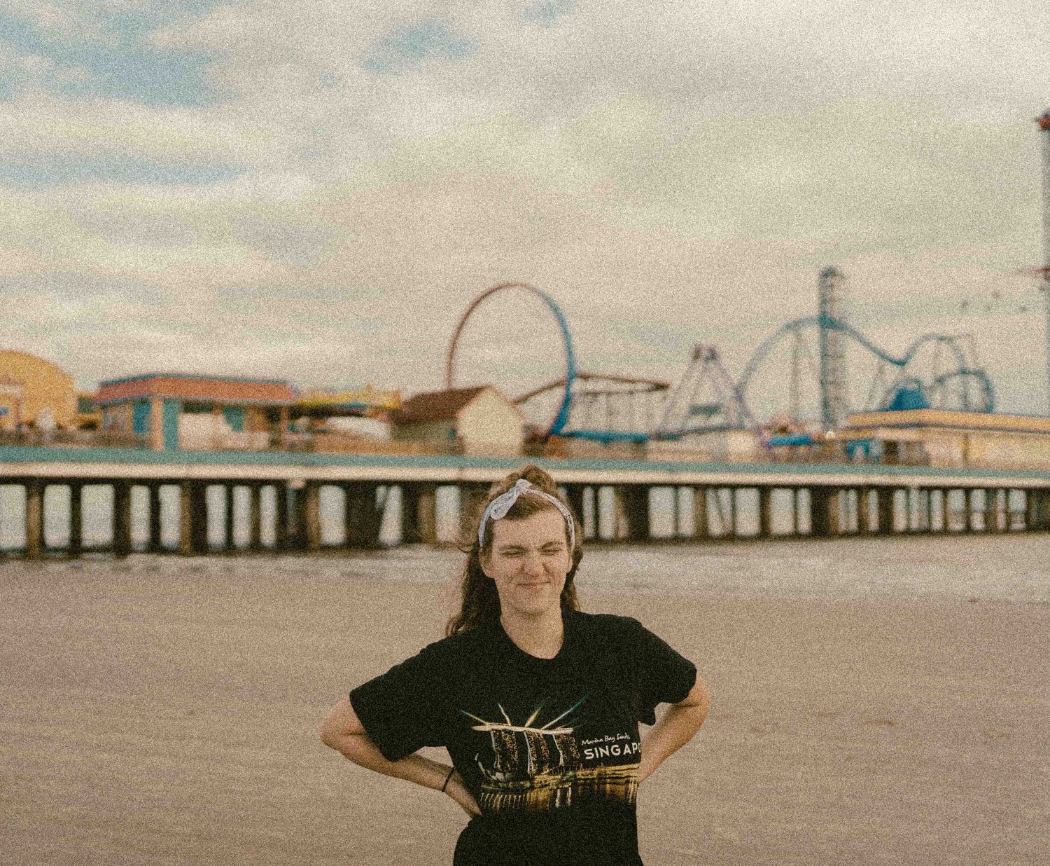 Person smiling on a beach boardwalk with colorful amusement park rides and a pier in the background.