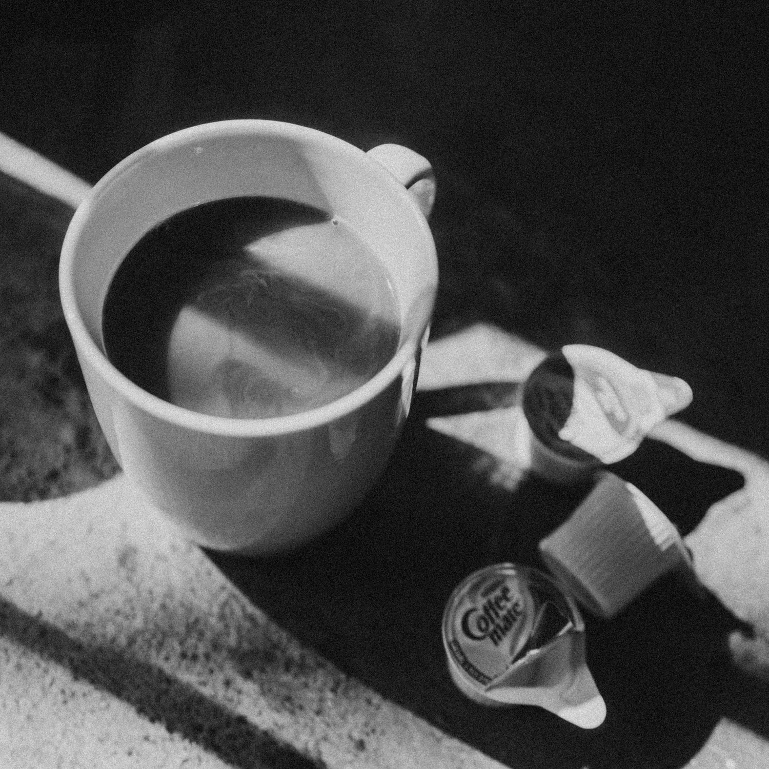 Black and white photo of a white coffee mug filled with dark coffee beside an open Coffee Mate creamer cup.