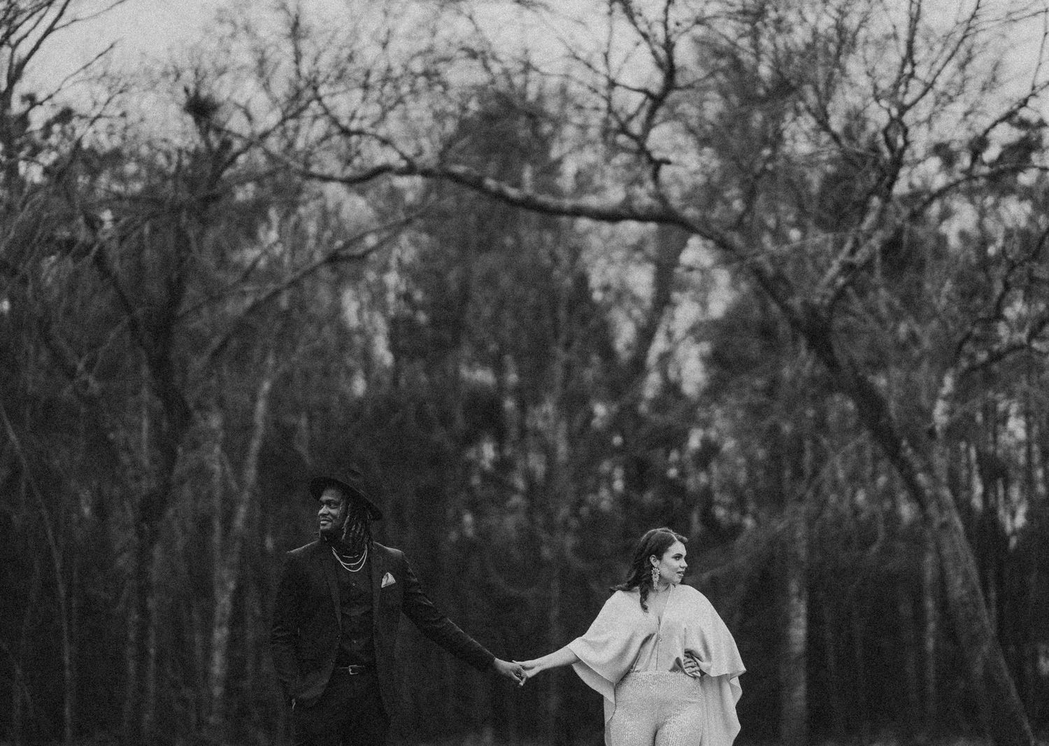 Black and white photo of a couple holding hands walking through a forest in wedding attire.