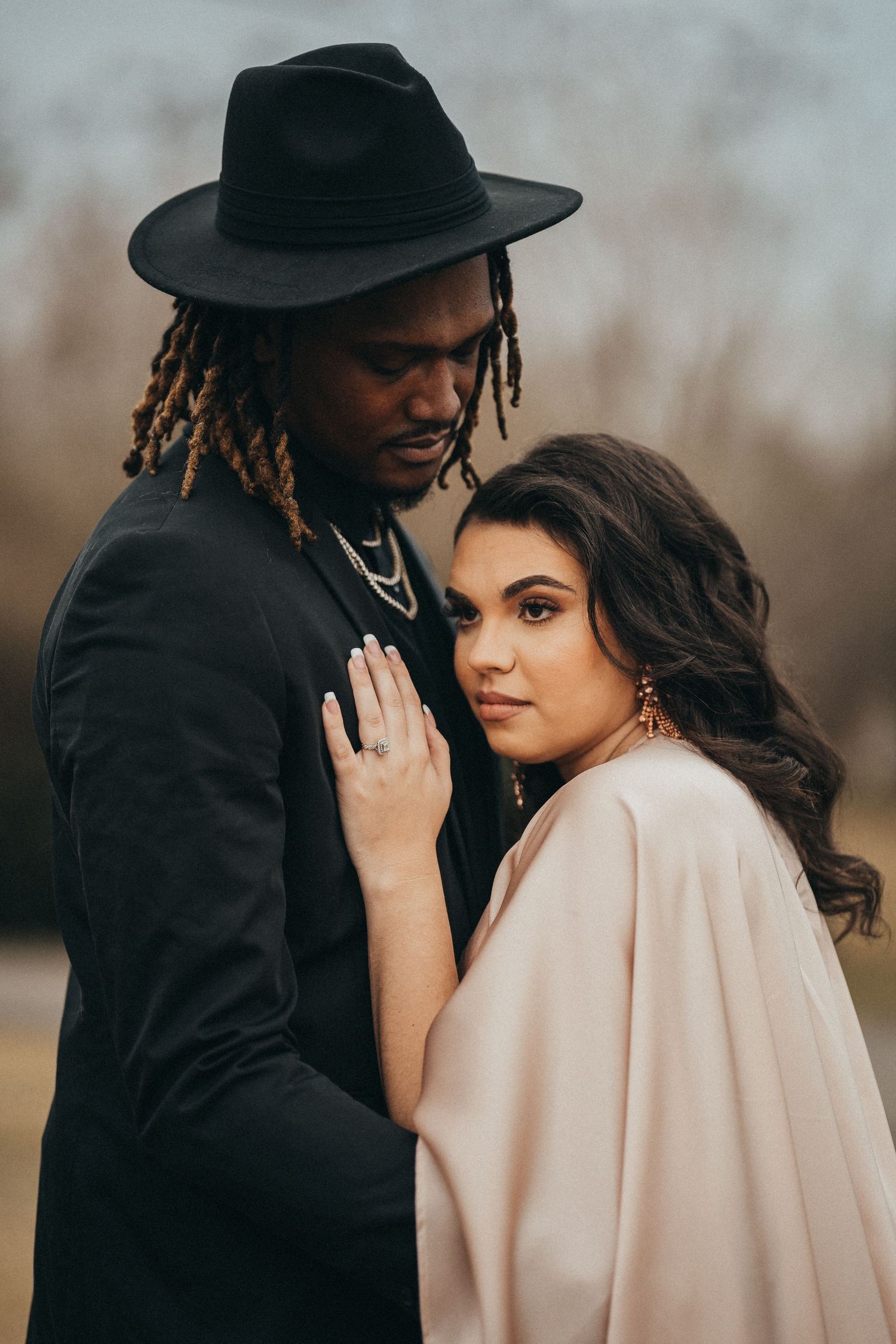 An elegant interracial couple poses outdoors, man in black suit and hat, woman in blush wrap.