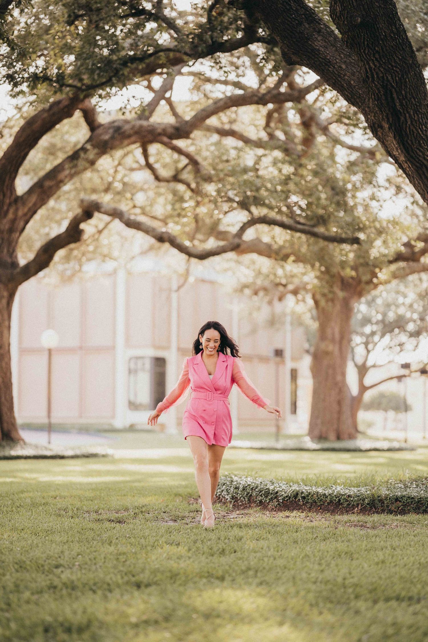 Woman in pink dress walking barefoot on green grass under large oak trees in a sunny park setting.