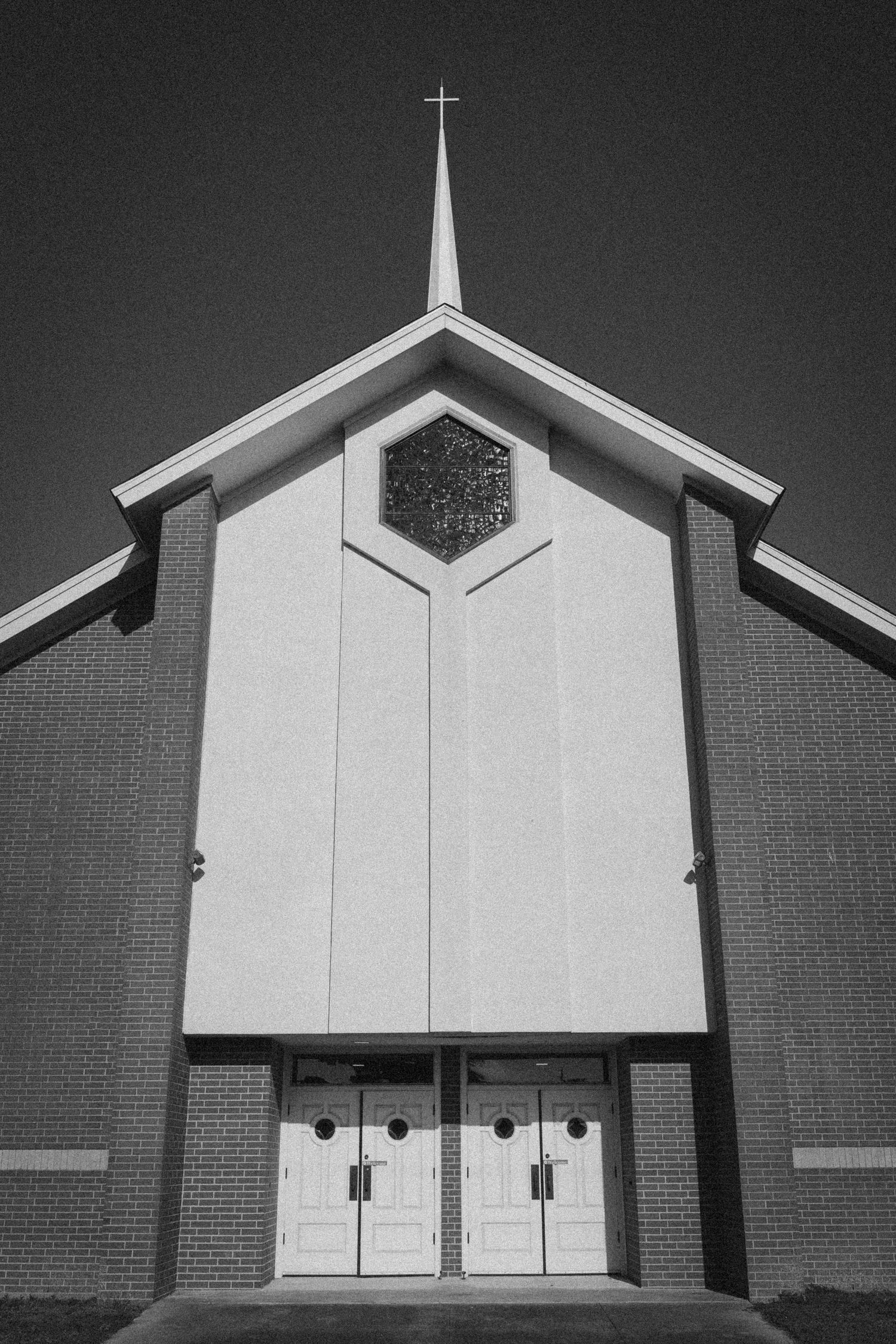 Black and white photo of a modern Christian church facade with steeple, cross, hexagonal window, and white double doors.