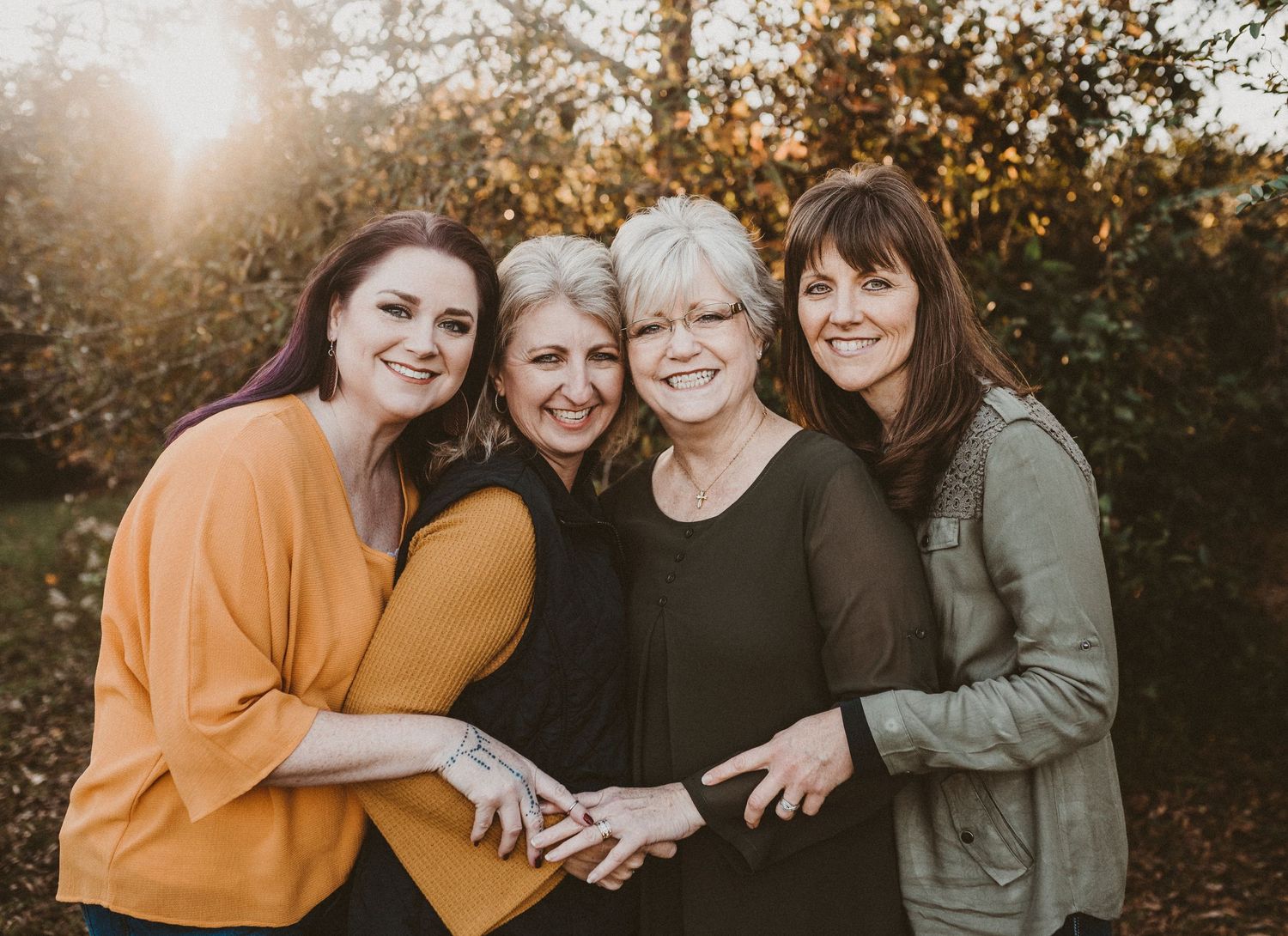 Four women smiling and hugging outdoors in golden sunlight among autumn foliage.