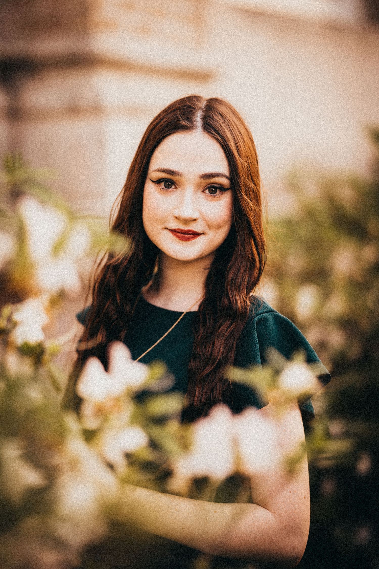 Young woman with auburn hair in green dress surrounded by white blooming flowers in a softly lit outdoor portrait.