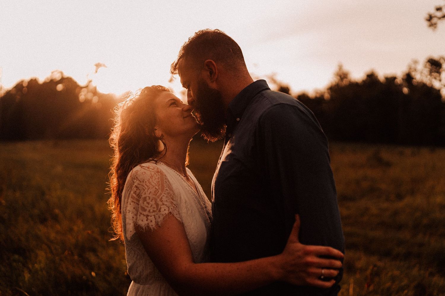 Couple sharing a romantic sunset kiss in a golden field, bride in lace dress embracing bearded groom.