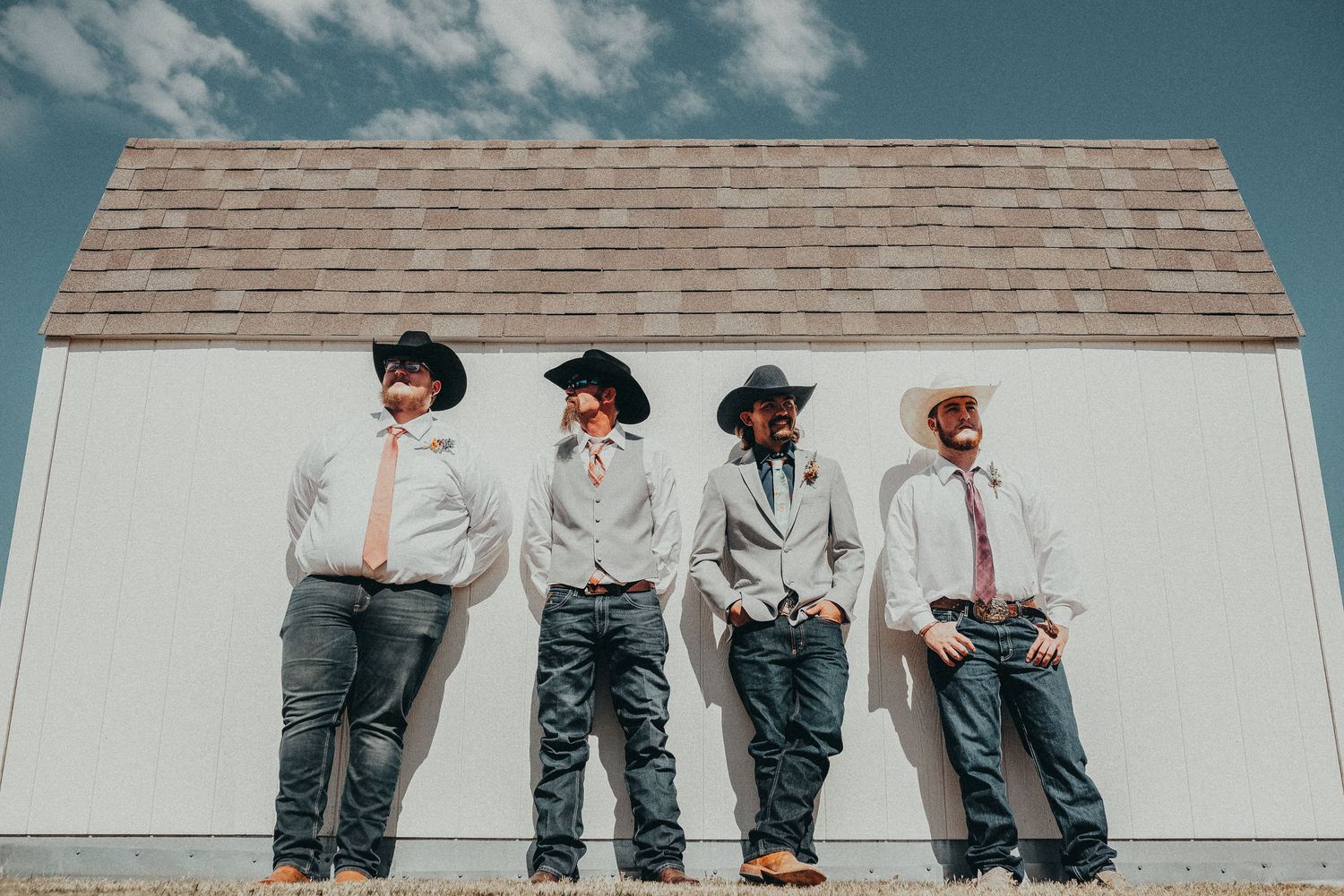 Four groomsmen in cowboy hats, dress shirts, and jeans pose against a white barn wall under a blue sky.