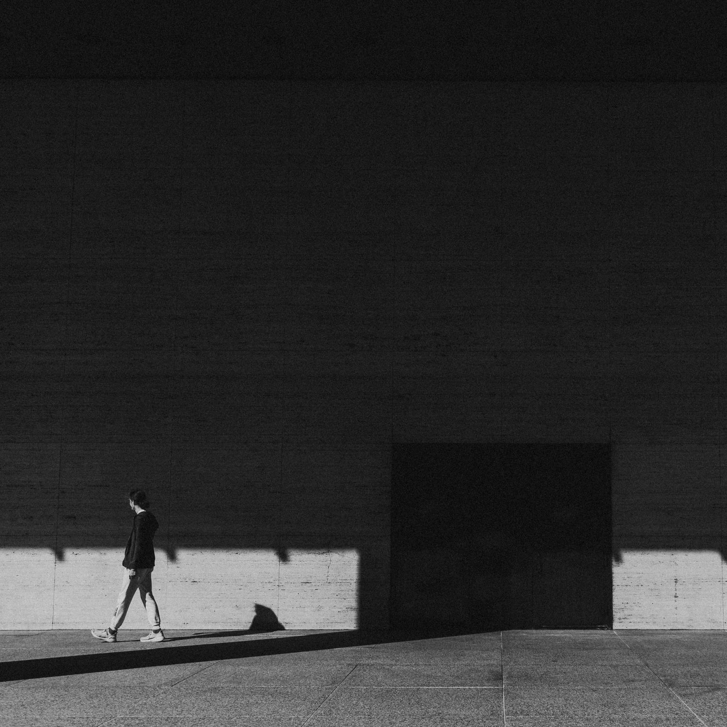 A lone person walks past a stark brick wall casting dramatic shadows in a minimalist black and white urban photograph.