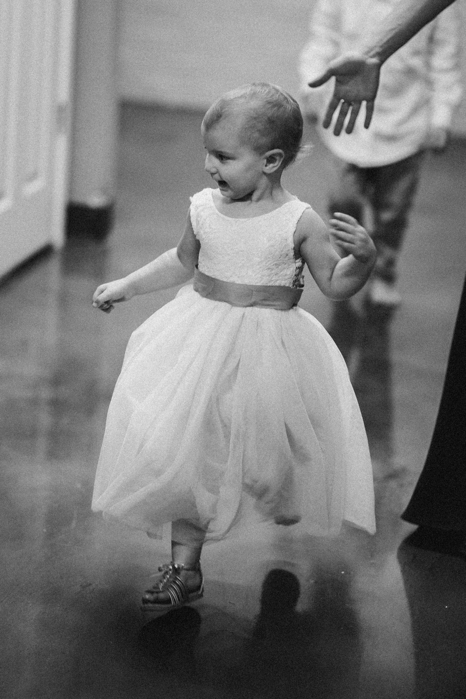 Adorable toddler flower girl in white tulle dress walking during wedding ceremony in black and white photo.