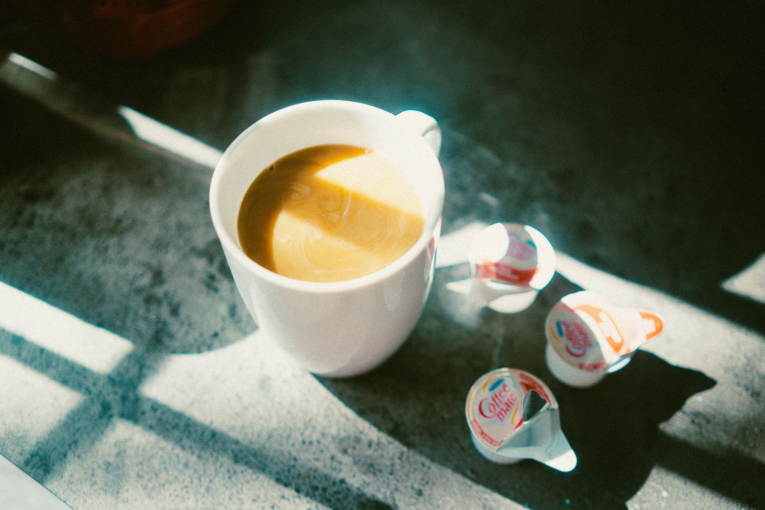 White ceramic cup of espresso coffee with creamer pods on a textured surface in bright sunlight.