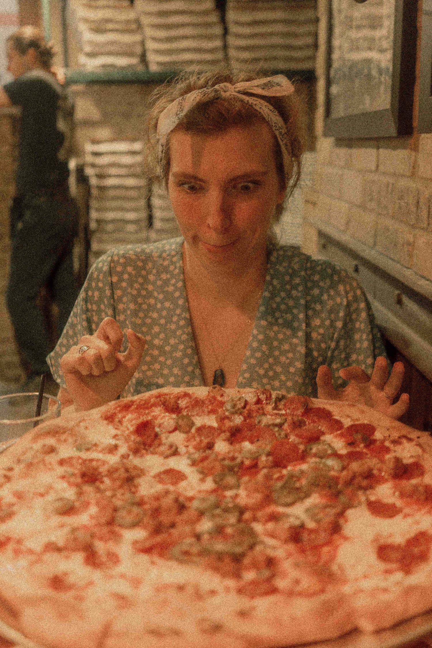 A woman with a headband sits eagerly before a large pepperoni pizza in a cozy brick-walled restaurant.