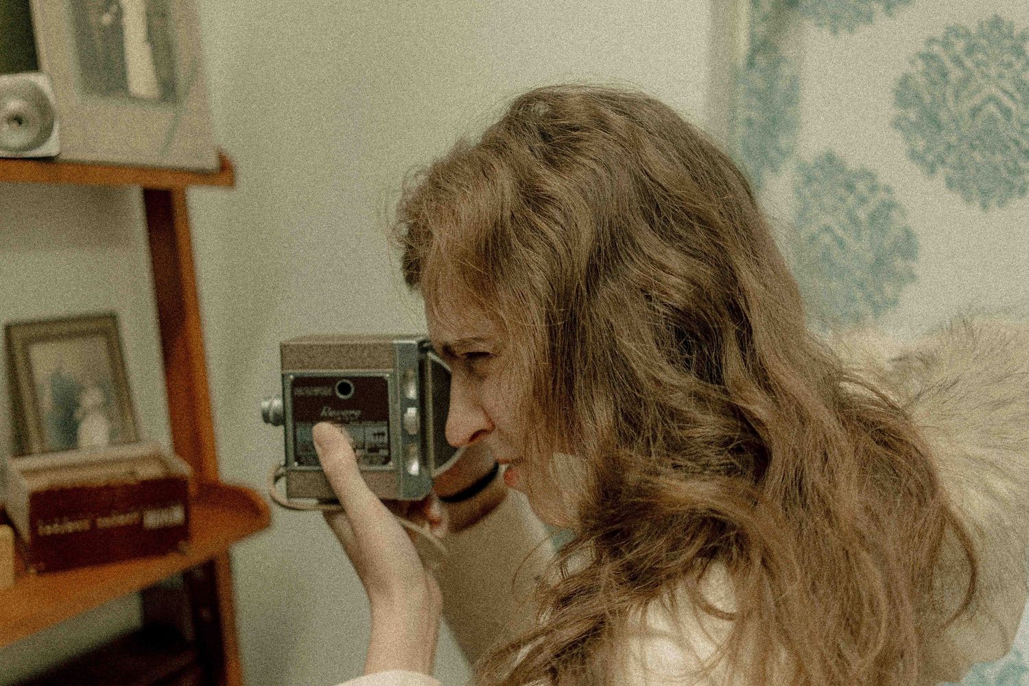 A young woman with long wavy hair holds a vintage camera up to her eye in a retro-styled room.