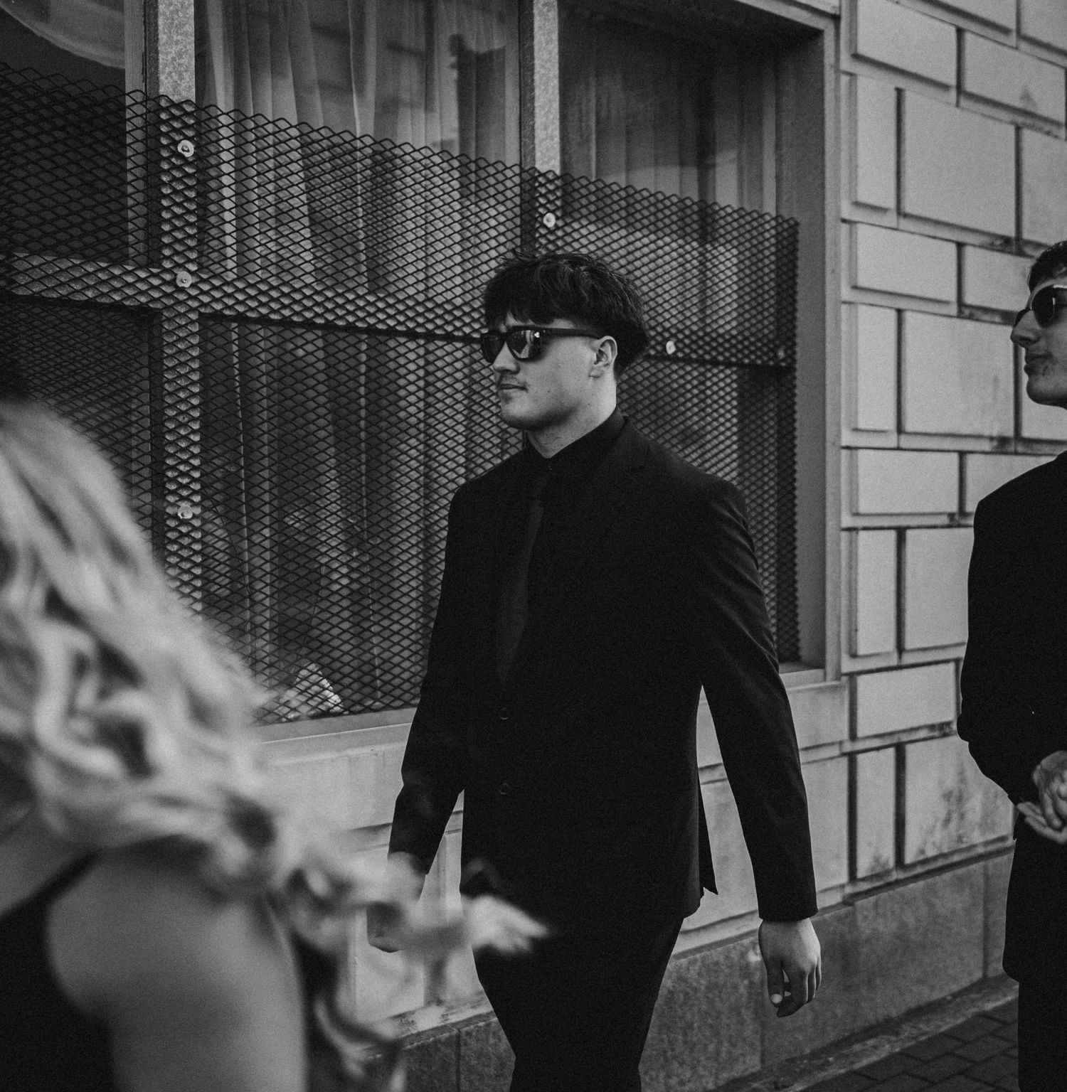 Man in black suit and sunglasses walking outdoors in black and white urban street photography.