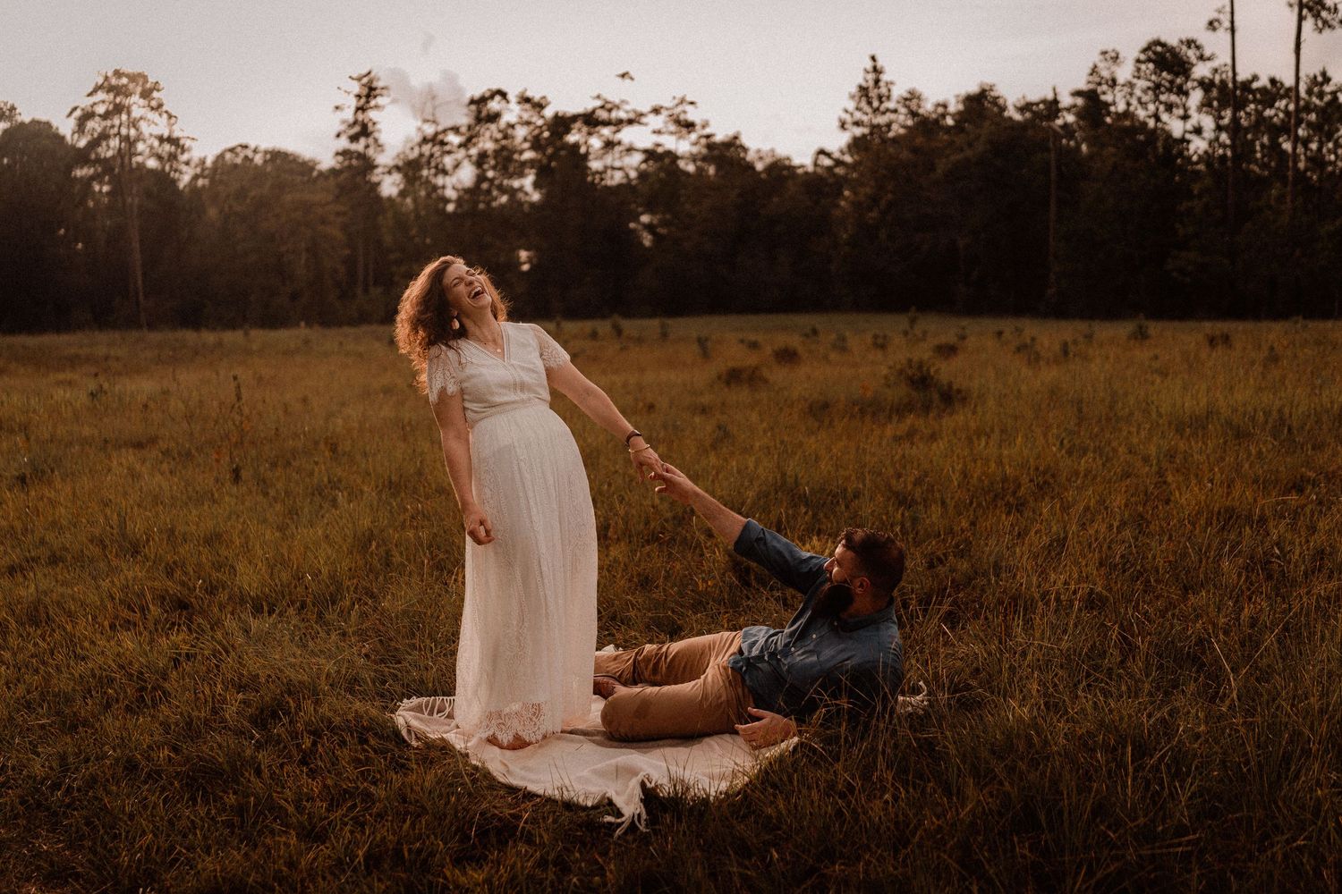 Pregnant woman in white dress holds hands with man sitting on blanket in golden hour field.