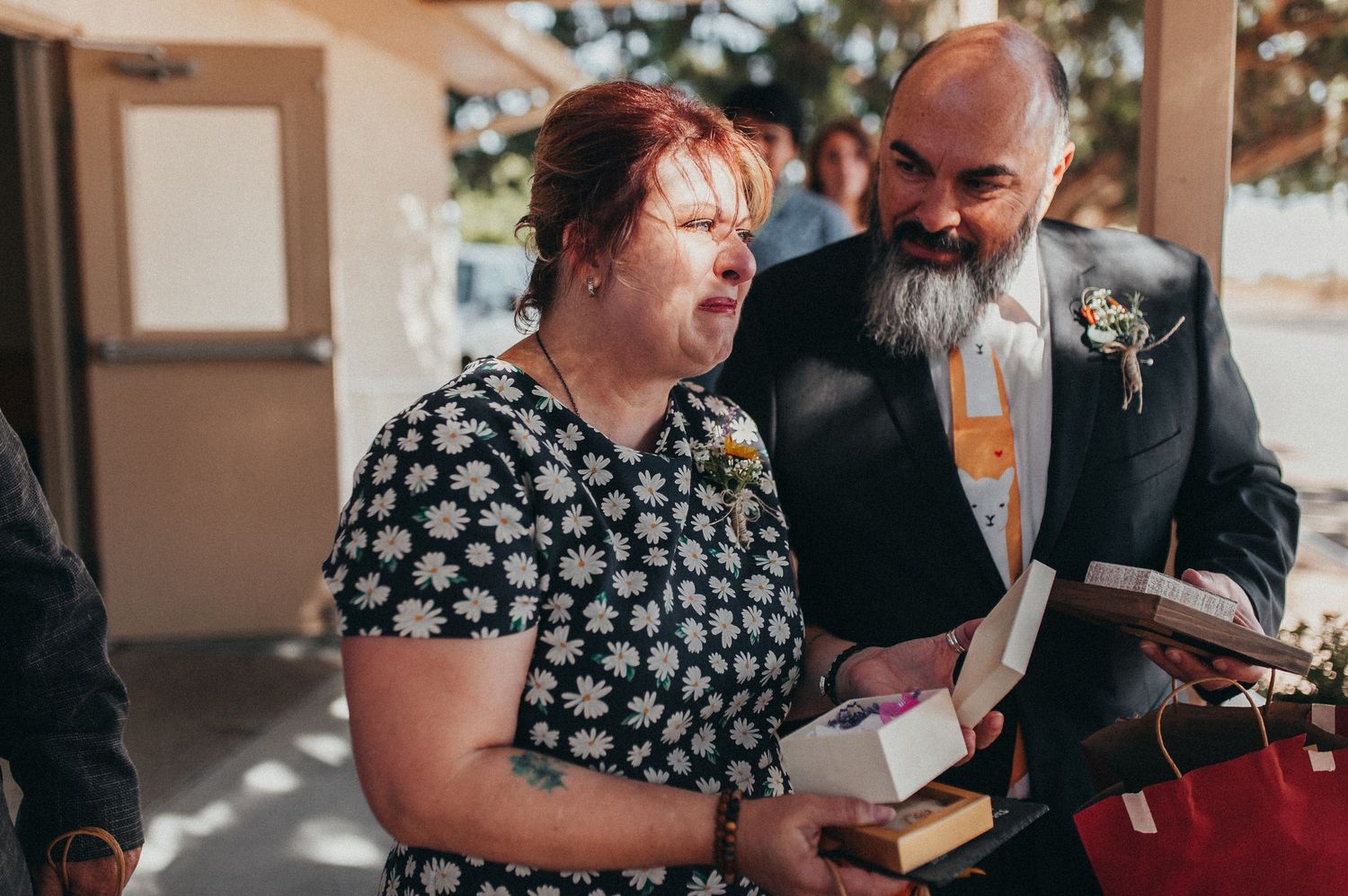 A woman in a floral dress and a bearded man in a suit share a joyful moment at a wedding reception.