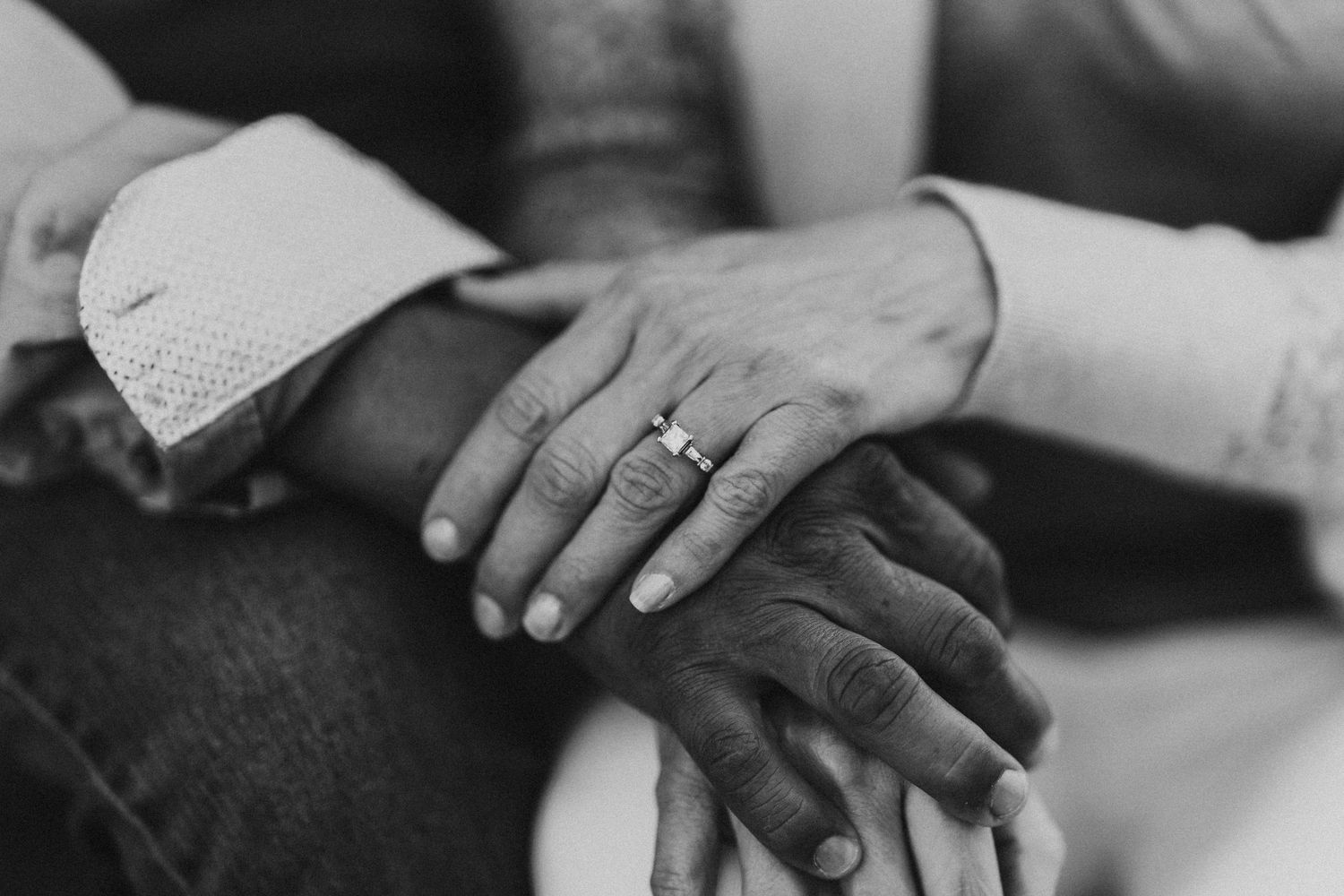 Black and white photo of hands stacked together, one wearing a diamond engagement ring, symbolizing love and commitment.