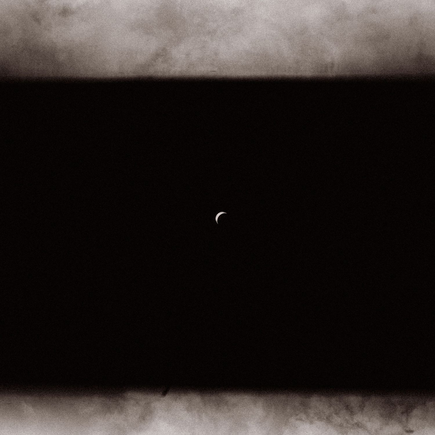 Dark moody photograph of a crescent moon barely visible through thick storm clouds in night sky.