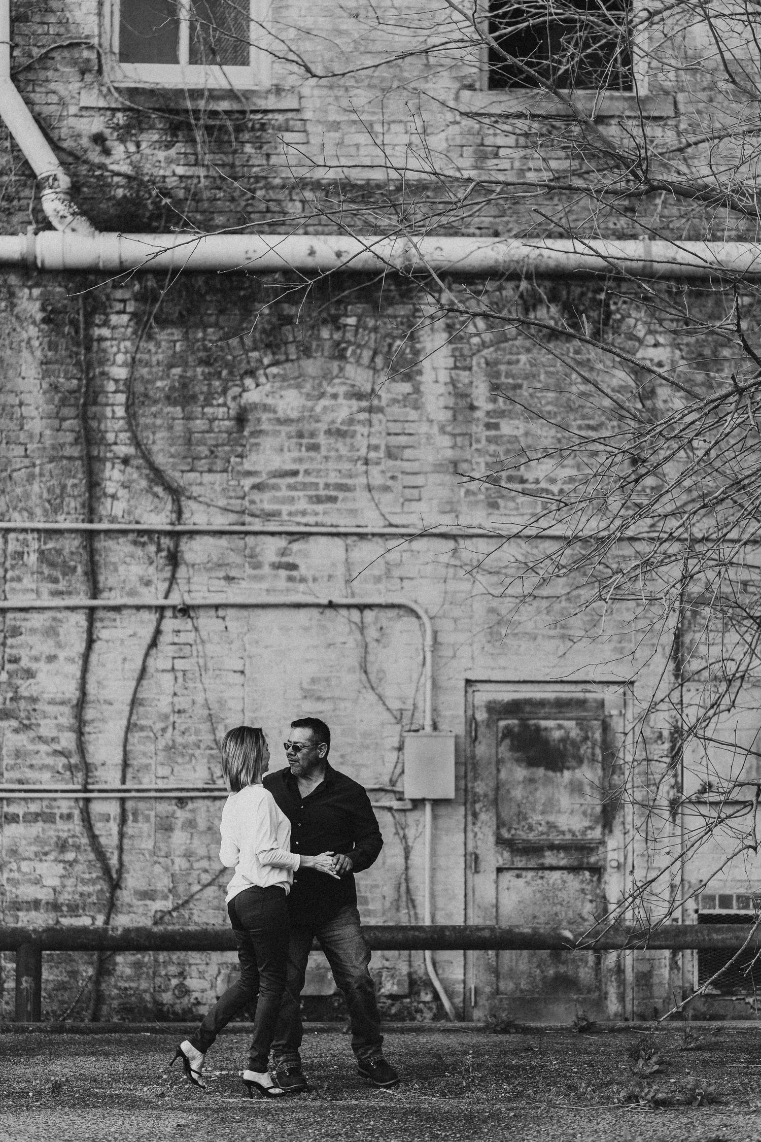 Black and white photo of a couple dancing playfully outside an old brick building.