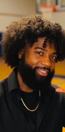 Young Black man with natural afro hair and beard, wearing black shirt and gold chain necklace, smiling warmly.