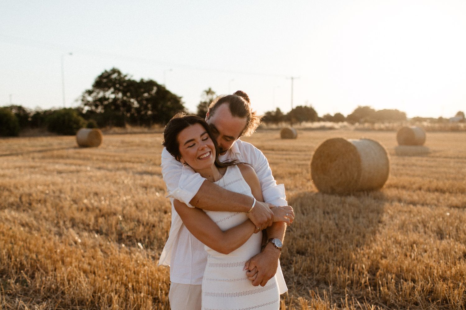 Couple embracing in golden wheat field with hay bales at sunset, wearing white outfits in romantic outdoor photo.