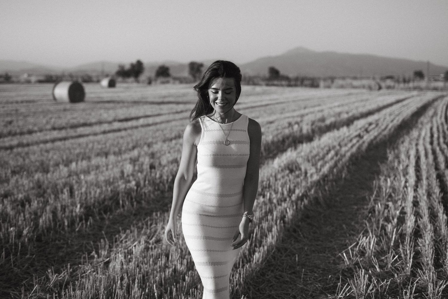 Black and white photo of a woman in a fitted dress walking through a harvested field in Larisa, Greece