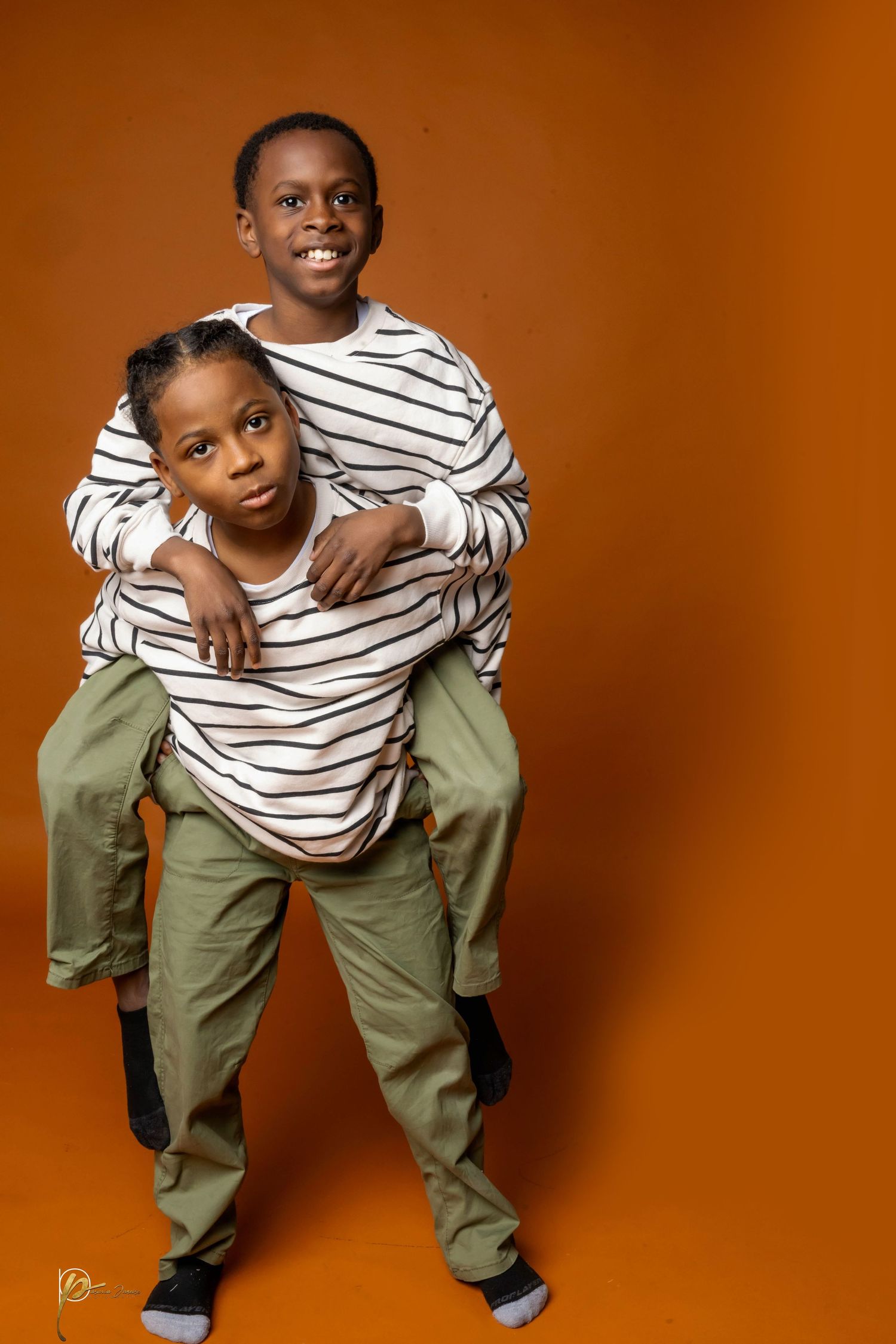 Two young boys in matching striped shirts and green pants pose together against an orange background.