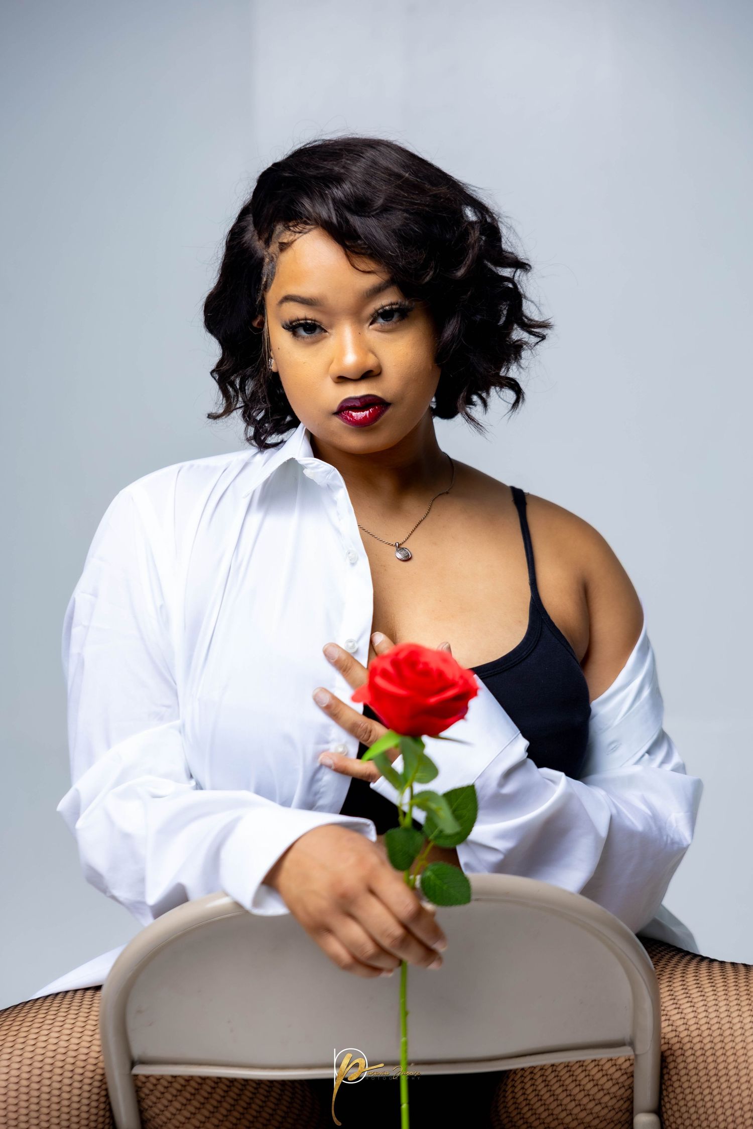 Young Black woman with short curly hair holds a red rose while seated, wearing a white shirt over a black top.